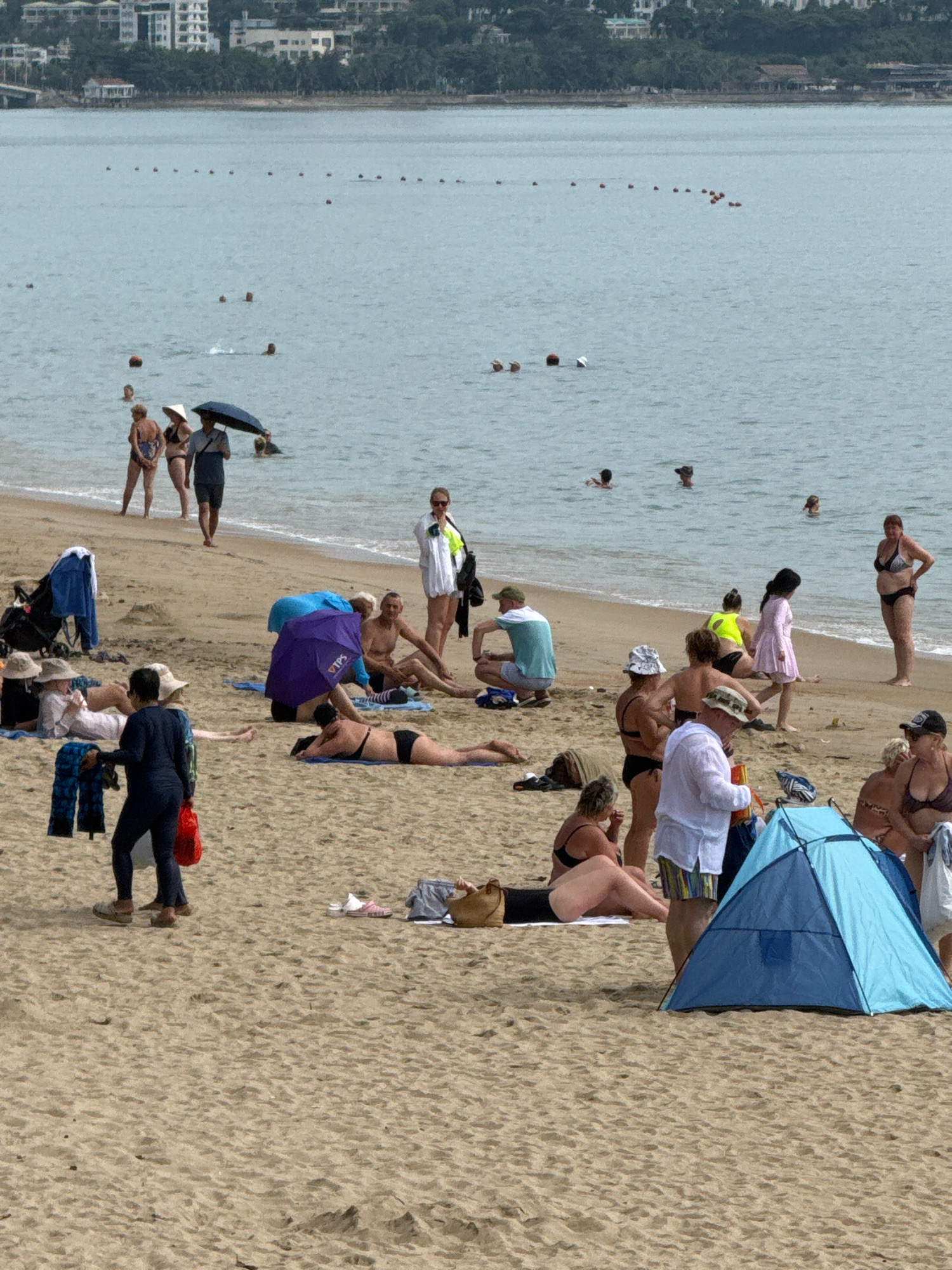 This is an eye-level, medium shot of a crowded beach in Nha Trang, Vietnam, likely on a warm, overcast day. The sand is a light brown, with ripples and footprints indicating recent activity. In the background, the sea is a muted blue-green, dotted with a few buoys. A distant shoreline is lined with buildings, some appearing to be hotels, and lush green trees.
The beach is populated with numerous individuals engaged in various activities. In the foreground, a small, bright blue beach tent is set up. A person in a white shirt and striped shorts stands near the tent. To the left, a woman in a black bikini is lying on a towel. Further back, several people are seated or reclining on the sand, some under umbrellas. One woman, wearing a yellow top and black bottoms, stands near the water's edge.
Further up the beach, a group of people are walking along the shoreline. One man, holding a dark umbrella, is accompanied by two women in bikinis. A stroller is parked near a woman in a white hat. Other individuals are scattered throughout the scene, some appearing to be wading in the shallow water. The overall impression is a lively, relaxed beach scene with a mix of locals and tourists enjoying the seaside.