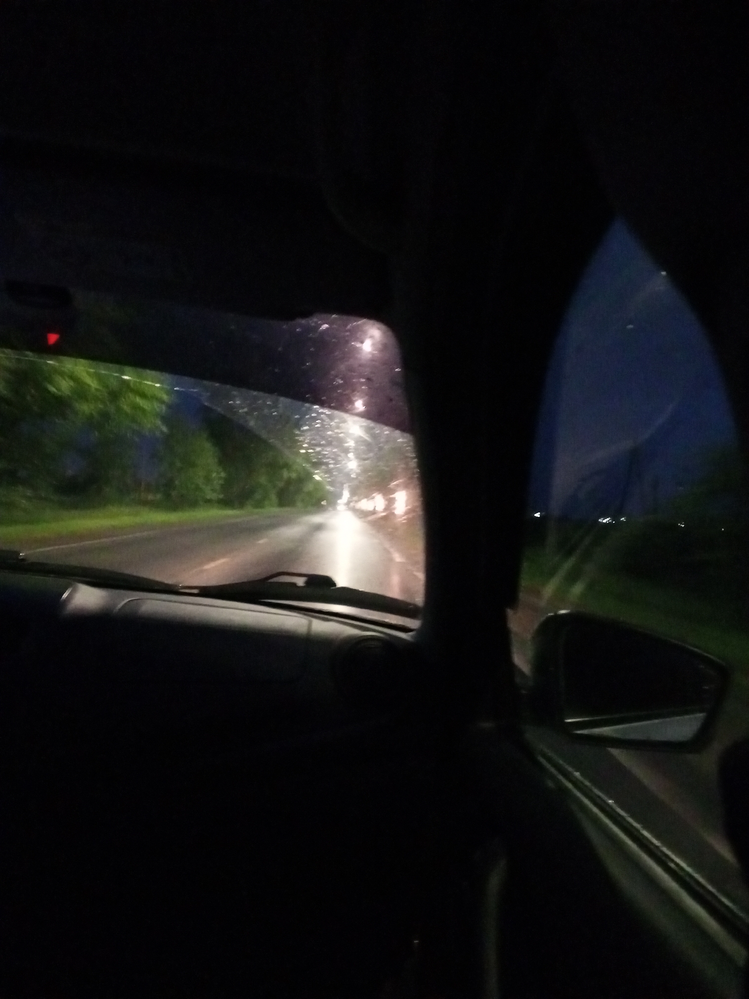 The image depicts a view from inside a car at night, looking out through the windshield. The scene shows a road stretching into the distance, lined with trees on the left. The road appears wet, with reflections of lights visible, suggesting it might be raining or has recently rained. Streetlights or vehicle headlights illuminate the road ahead, creating a blurred, streaky effect due to the motion and possibly the wetness on the windshield.
The interior of the car is mostly dark, with the dashboard and parts of the car's structure visible in silhouette. A small red light is visible on the upper left part of the windshield area. The overall impression is one of driving at night, possibly in damp conditions. The time of day is clearly night, indicated by the darkness and artificial lights. There are no people clearly visible, but the perspective suggests a person is driving or being driven. The setting appears to be a road through a wooded or vegetated area. The mood evoked is one of quiet travel, with the ambient light and reflections creating a somewhat atmospheric or contemplative feel. No text is visible in the image.