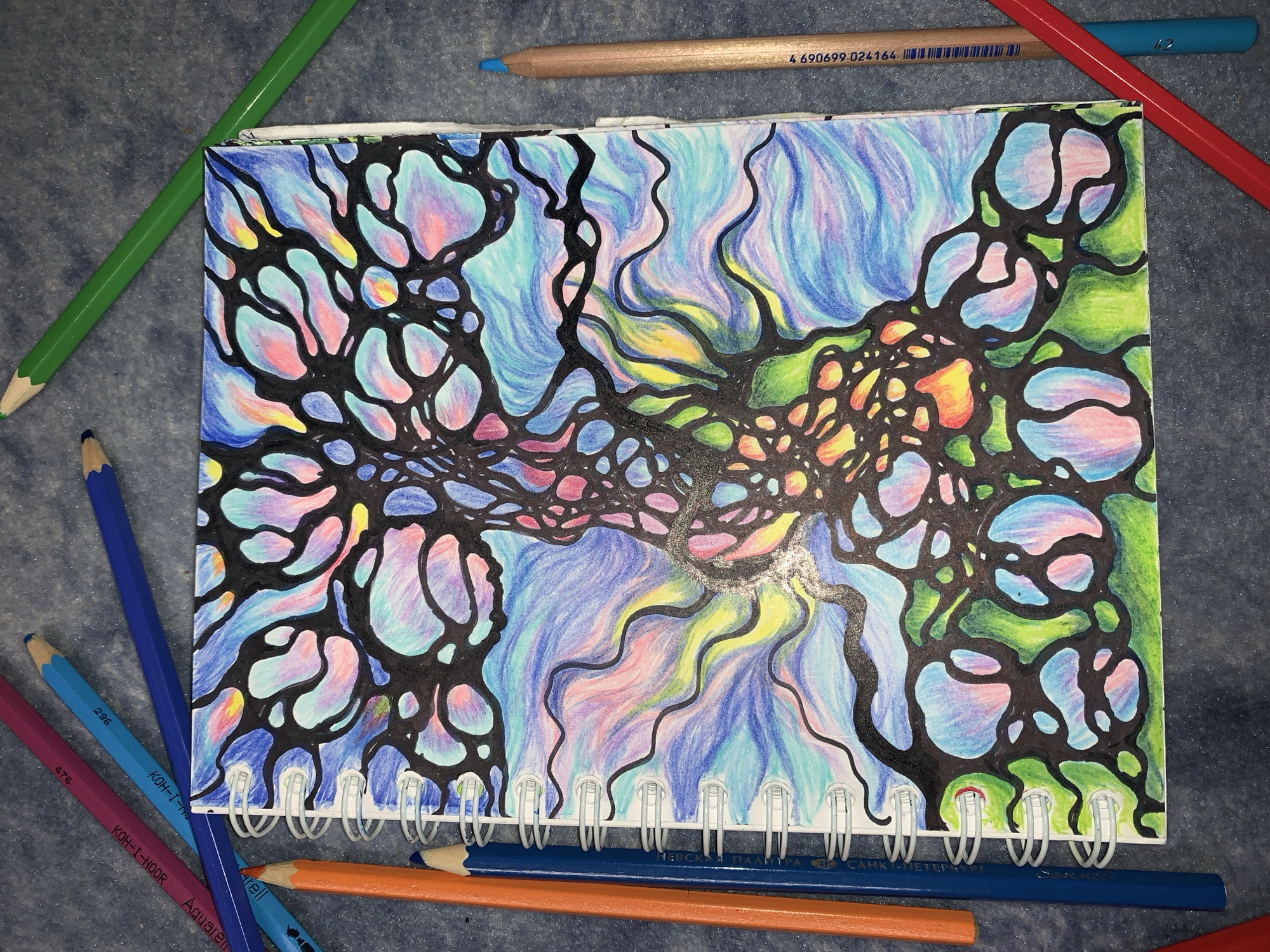 The image shows a notebook with a colorful abstract drawing. The drawing is done in a style that resembles neural networks or abstract organic shapes, with black outlines filled with vibrant colors like blue, green, pink, yellow, and orange. The background of the drawing features flowing lines in shades of blue and purple, creating a sense of movement.
The notebook is spiral-bound and lies open on a dark, textured surface, possibly fabric or a dark countertop. Several colored pencils are arranged around the notebook, including green, red, blue, orange, purple, and a light brown one. Some of the pencils have visible text printed on them, such as "KOH-I-NOOR" and what appears to be a barcode and numbers "4 690699 024164" on the light brown pencil. Other text fragments are partially visible on some pencils. The lighting suggests a well-lit indoor environment. No people are present in the image. The overall impression is one of creativity and artistic exploration.