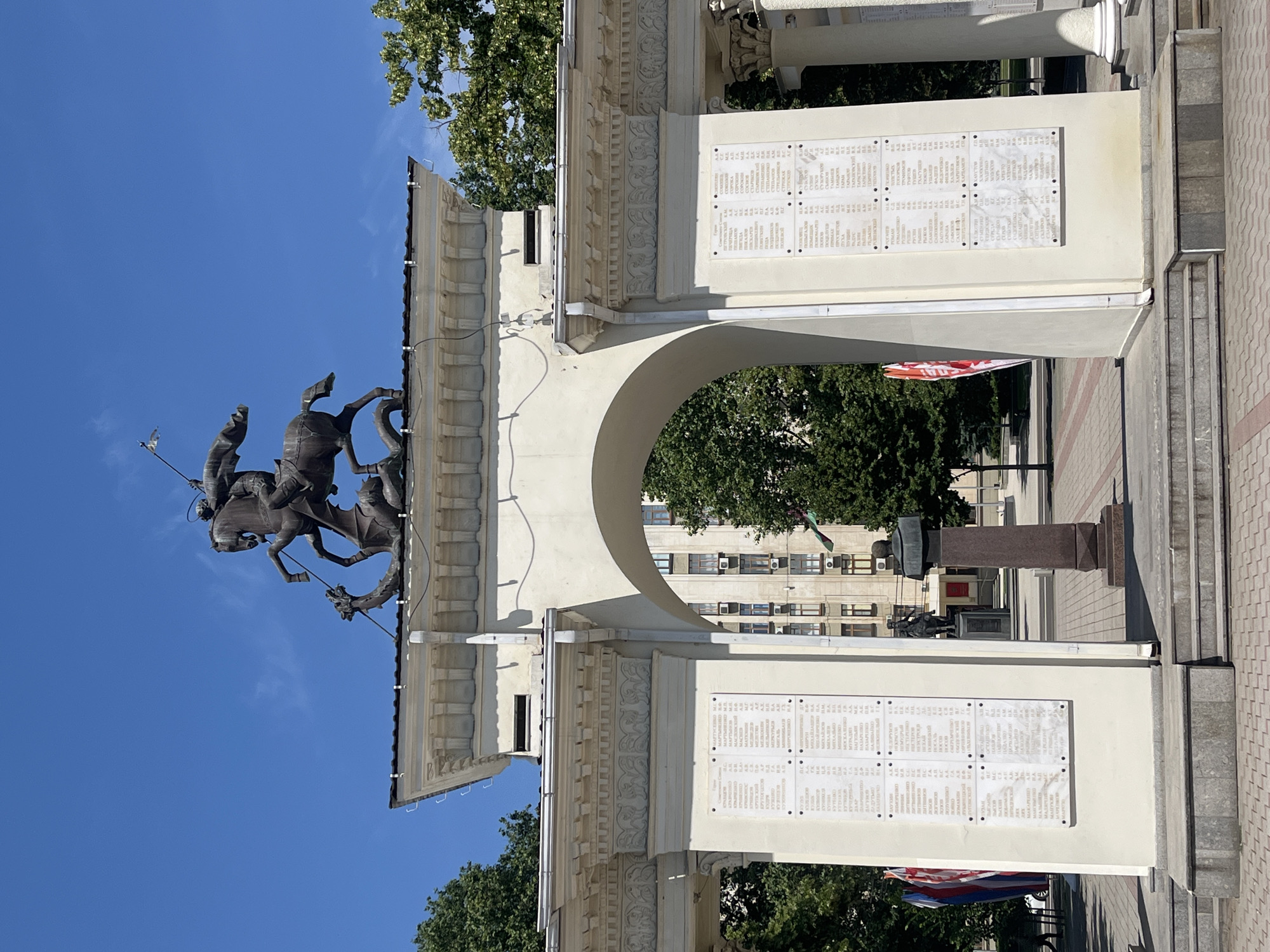 The image shows a monument dedicated to the heroes of the Great Patriotic War in Krasnodar, Russia. The monument features a bronze sculpture of Saint George slaying a dragon, mounted on top of a large archway. The archway is flanked by two columns and has white stone panels on either side, inscribed with lists of names. A statue of a soldier is visible in the background, along with a building and trees. The sky is clear and blue.
