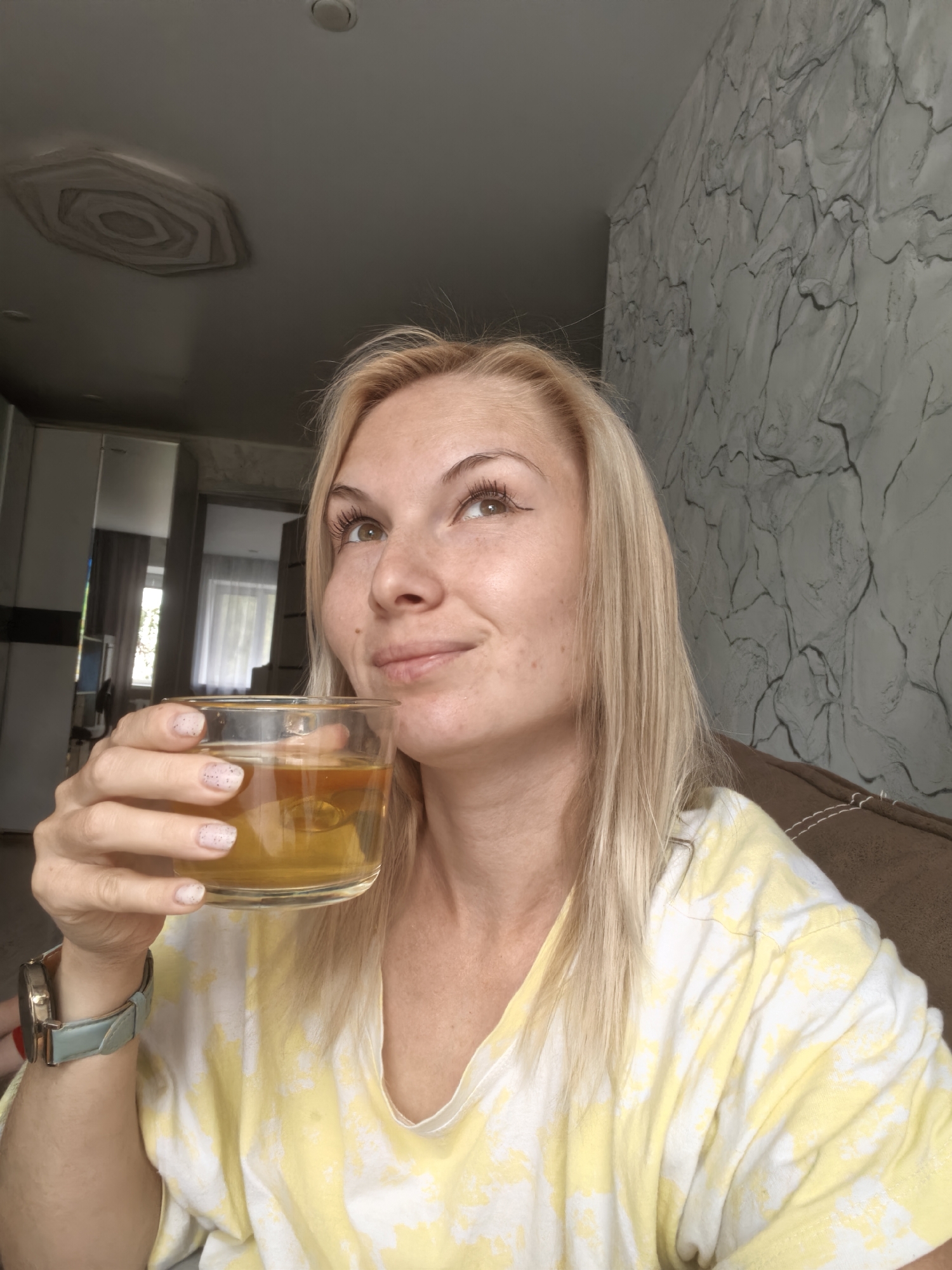 A woman with blonde hair and light skin is holding a glass of amber liquid. She is wearing a yellow and white tie-dye t-shirt and a light blue watch on her left wrist. Her eyes are looking up and to the right. The background shows a wall with a marble-like pattern and a section of the ceiling. The image appears to be taken indoors. The location is not specified as Sterlitamak, Russia.