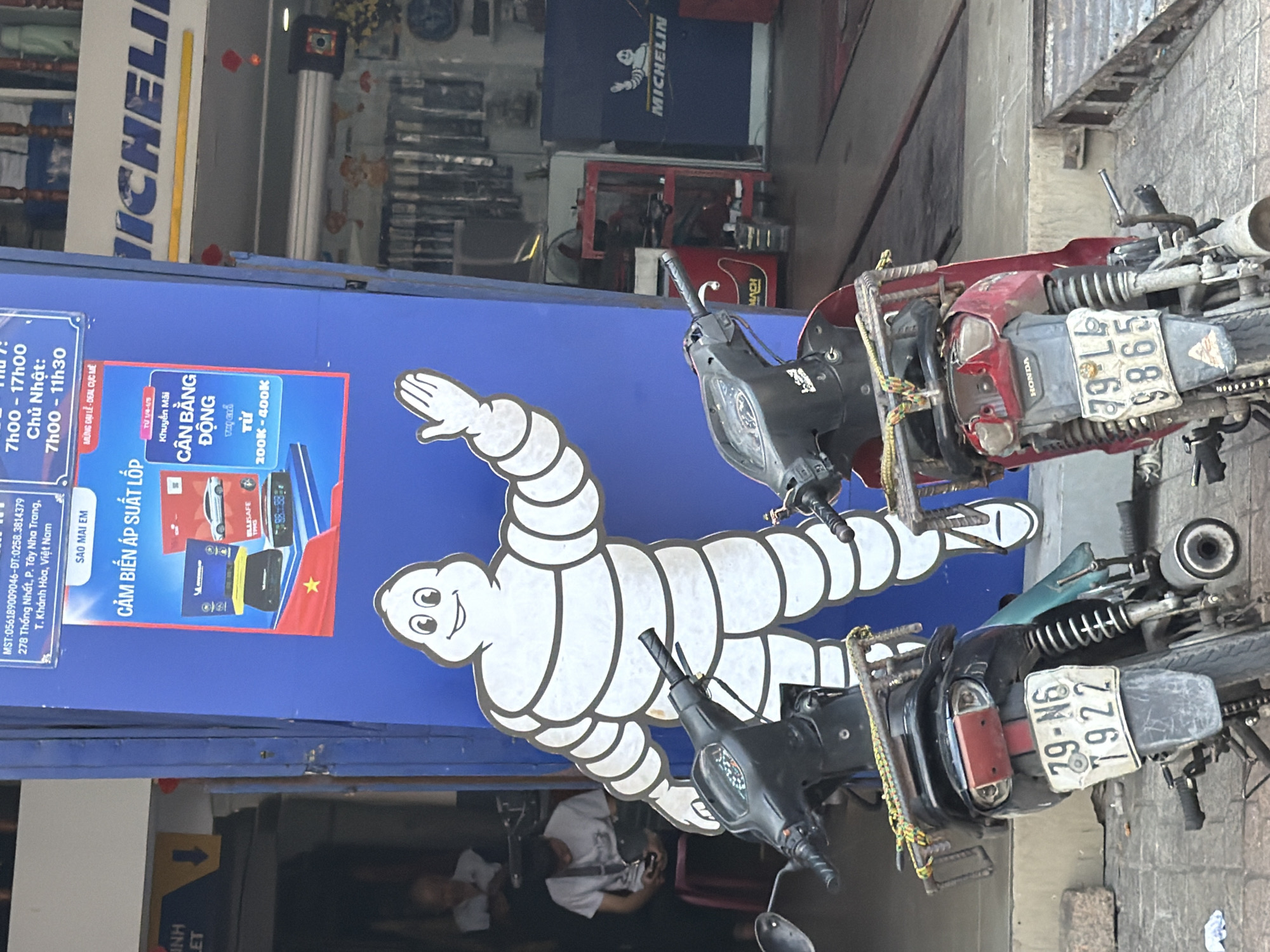 A street scene in Tay Nha Trang Ward, Vietnam, features a blue building with a large Michelin Man cutout and a poster advertising tire pressure sensors. Two motorcycles are parked in front of the building. The poster displays the text "SAO MAI EM" and "CAM BIEN AP SUAT LOP," along with product images and pricing information. The building's signage includes "MICHELIN." Inside the building, shelves stocked with automotive supplies are visible. In the background, several people are seated, and one individual is looking at a mobile phone. The environment appears to be an automotive service or retail establishment.