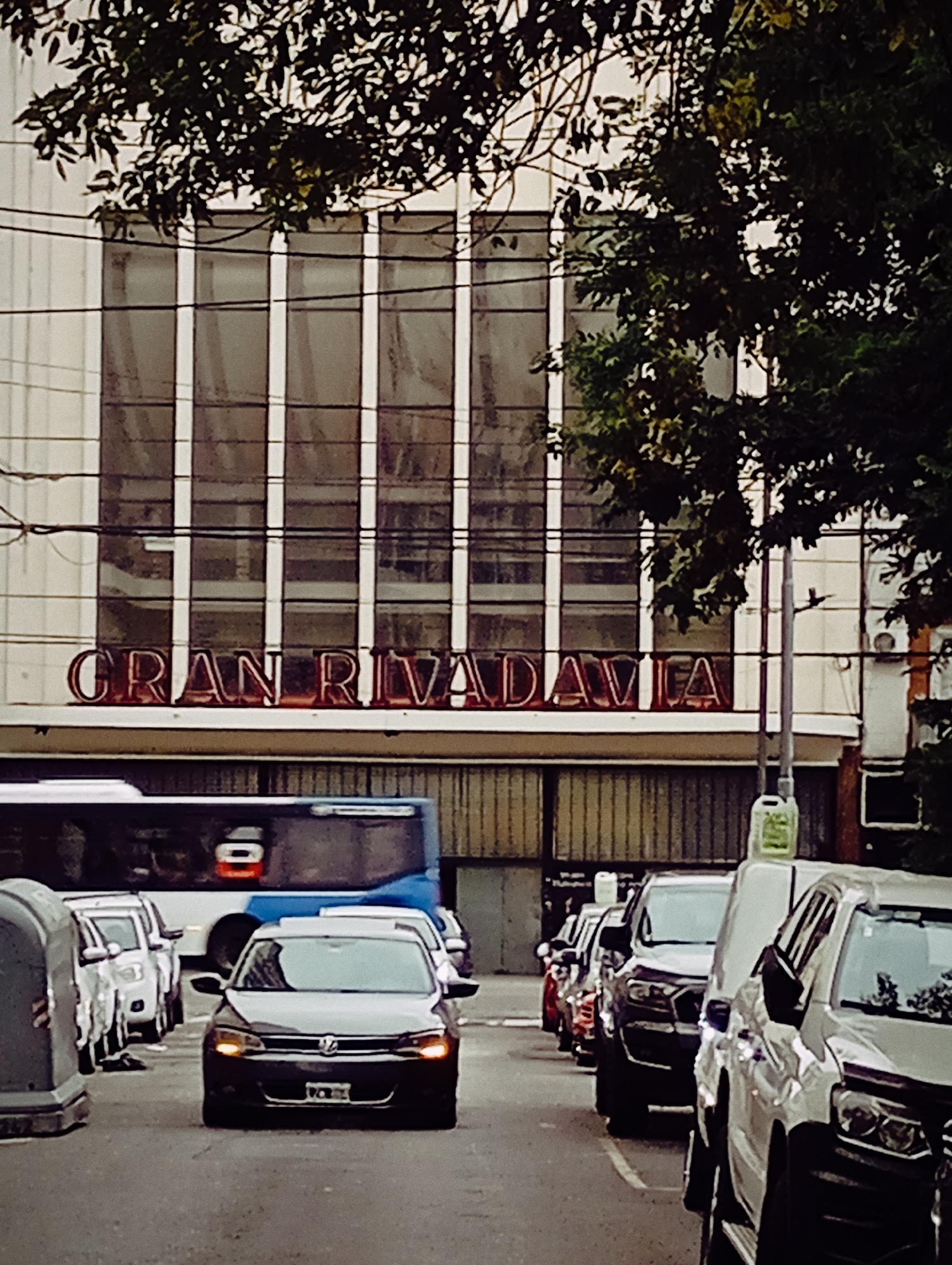 The image shows a street scene in Buenos Aires, Argentina, in front of the "Gran Rivadavia" building. A line of parked cars occupies the right side of the street, while a black Volkswagen sedan is driving in the center lane. Behind the Volkswagen, a bus is partially visible. The "Gran Rivadavia" building features a white facade with large, rectangular windows. Trees partially obscure the building at the top. Overhead power lines cross in front of the building.