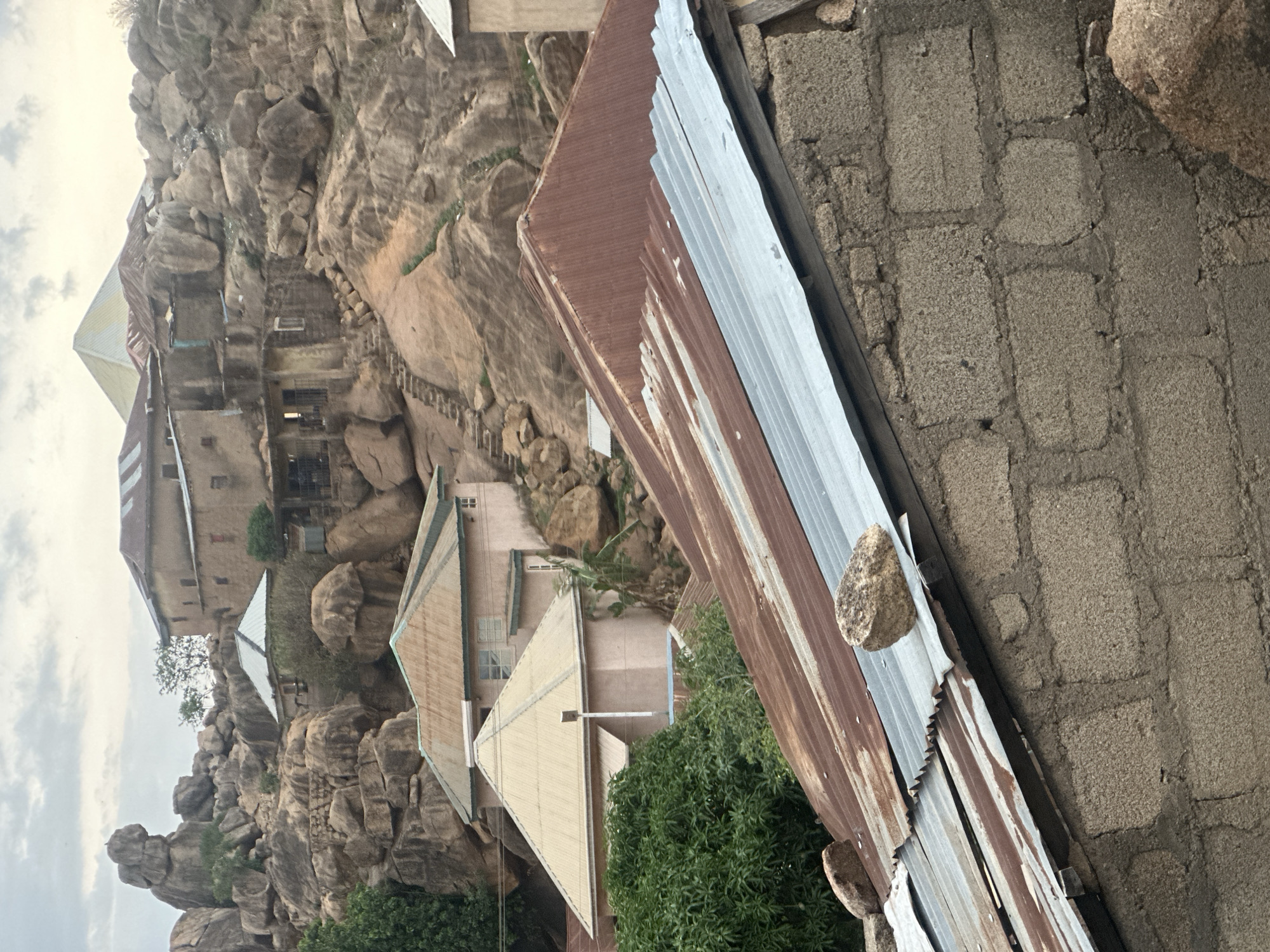 The image shows a view of buildings nestled among rocky hillsides in Jos, Nigeria. The foreground contains a partially collapsed corrugated metal roof leaning against a rough, brick-like wall. The background reveals multiple buildings of varying heights and colors (pink, beige, tan) interspersed among large rock formations and sparse vegetation. The roofs of these buildings are predominantly peaked. A set of rough stairs is visible, cut into the rock face. The sky is overcast.