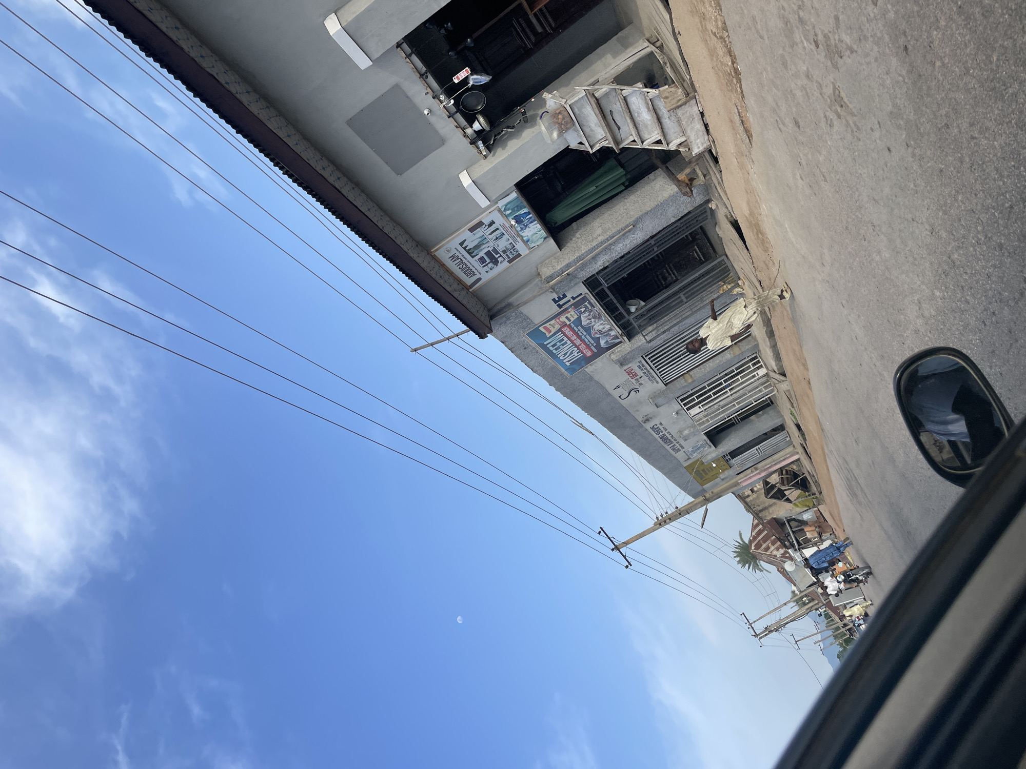The image captures a street scene in Jalingo, Nigeria, oriented vertically. In the foreground, a partial view of a car's interior, specifically a side-view mirror and part of the dashboard, suggests the photo was taken from a moving vehicle.
The main subject is a row of shops lining a street. The buildings have a mix of modern and older architectural elements, with some featuring metal grates over windows and doorways. One building appears to be under construction or renovation, with visible scaffolding and building materials. Various signs are displayed on the storefronts. One sign, partially visible, reads "ABDULSALAM". Another sign has text in a foreign script, possibly Arabic, and an image of people. A motorbike is parked in front of one of the shops.
A man in traditional light-colored clothing stands on the sidewalk in front of a shop. Further down the street, several other individuals can be seen, along with what appear to be more shops and a palm tree. The sky above is a clear, bright blue with scattered white clouds, indicating it is daytime, likely late morning or early afternoon. The moon is also faintly visible in the sky. Electrical wires crisscross the scene, supported by wooden poles, a common sight in many urban and semi-urban areas. The overall impression is of a busy, everyday street scene in a Nigerian town.