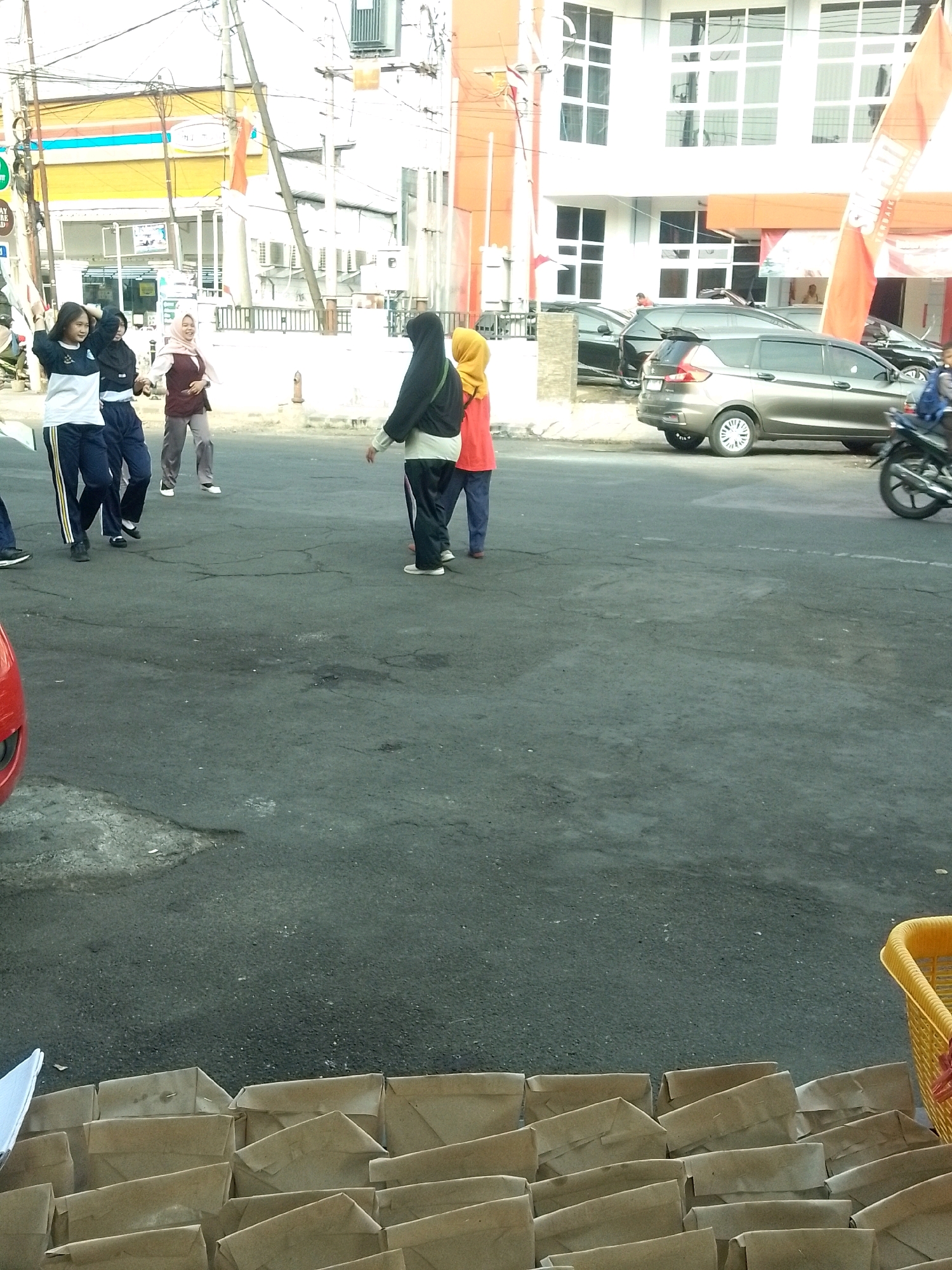 The image captures a street scene in Jember, Indonesia, likely during daylight hours given the bright ambient light. In the foreground, a row of brown paper bags is neatly stacked, suggesting a commercial or street vendor setup. A yellow plastic basket is visible to the right of the bags, also appearing to be part of the same setup.
Across the street, several individuals are present. On the left side of the road, a group of four young women are walking together. Three of them are wearing hijabs and athletic attire. The woman on the far left has long dark hair and is wearing a blue and white long-sleeved top with dark pants featuring yellow stripes. The woman next to her is wearing a black hijab and a similar outfit. The third woman is wearing a light pink hijab and a maroon top with grey pants. The fourth woman, on the right of this group, is wearing a black hijab and a black long-sleeved top with dark pants.
Further down the street, on the right side, two more individuals are visible. One person is wearing a yellow hijab and an orange shirt, and appears to be walking away from the camera. Another person in a black hijab and a white and black long-sleeved top with dark pants is also present.
Several vehicles are parked or driving on the street. A silver multi-purpose vehicle (MPV) is parked on the right side of the road. Further back, a black car is visible. A motorcycle is in motion on the far right of the image, with a rider wearing a blue jacket.
The background features buildings, including a white building with multiple windows and an orange accent. A prominent pole with electrical wires runs through the scene, a common sight in urban Indonesian environments. In the distance, there appears to be a yellow building with signage, possibly indicating a shop or business. The road surface is asphalt and shows some signs of wear and tear, with visible cracks.