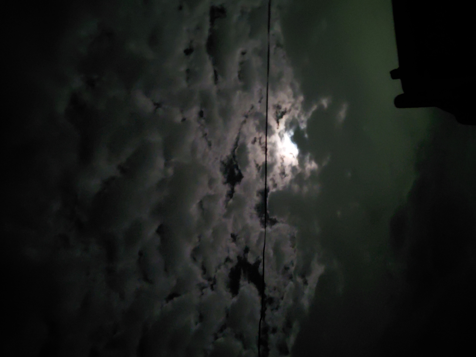The image displays a dark, cloudy night sky, with a bright celestial body, likely the moon, partially obscured by dense cloud formations. The sky transitions from deep blacks on the left to a dark greenish-gray hue on the right. The clouds are voluminous and textured, appearing in various shades of gray and white where illuminated by the light source, creating a dramatic, mottled effect across the frame.
A thin, dark vertical line, possibly a wire or cable, runs down the center of the image, cutting across the cloudy sky. In the upper right corner, a dark, silhouetted structure, which could be part of a building or roofline, is partially visible. The overall scene suggests a quiet, overcast night. No human activity or identifiable figures are present. The time of day is clearly night, characterized by the dark sky and the bright, diffused light from behind the clouds. There is no visible text in the image. This scene is situated in Santa Ana, Venezuela.