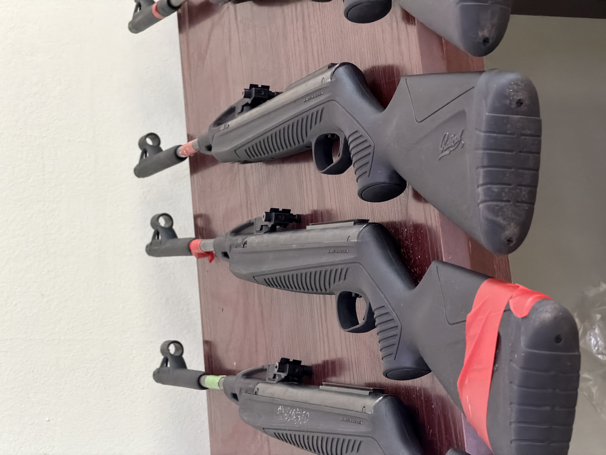 The image depicts a display of three air rifles mounted on a wooden panel. The rifles are gray with black accents and are secured in a vertical arrangement. Red and green tape is visible on the barrels and butts of the rifles. The background consists of a plain white wall and the wooden panel supporting the rifles. The location, Забегалово, Russia, cannot be determined from the provided image.