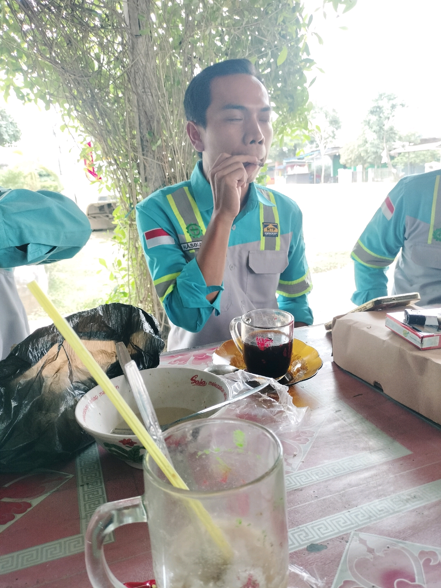 In the city of Maburai, Indonesia, two individuals in teal and grey work uniforms are seated at a red and white patterned table. The individual on the left is partially obscured by a blue fabric. The individual on the right, facing the camera, has their eyes closed and is bringing an object to their mouth. They wear a teal uniform with grey panels and reflective stripes, featuring a red and white Indonesian flag patch and a logo that reads "RTP GROUP". On the table, a glass cup filled with a dark beverage sits on a yellow saucer, next to a white bowl containing food and a yellow straw. A black plastic bag and other items, including a pack of cigarettes, are also visible. The background shows a natural setting with trees and foliage, along with a hint of buildings and what appears to be a vehicle.