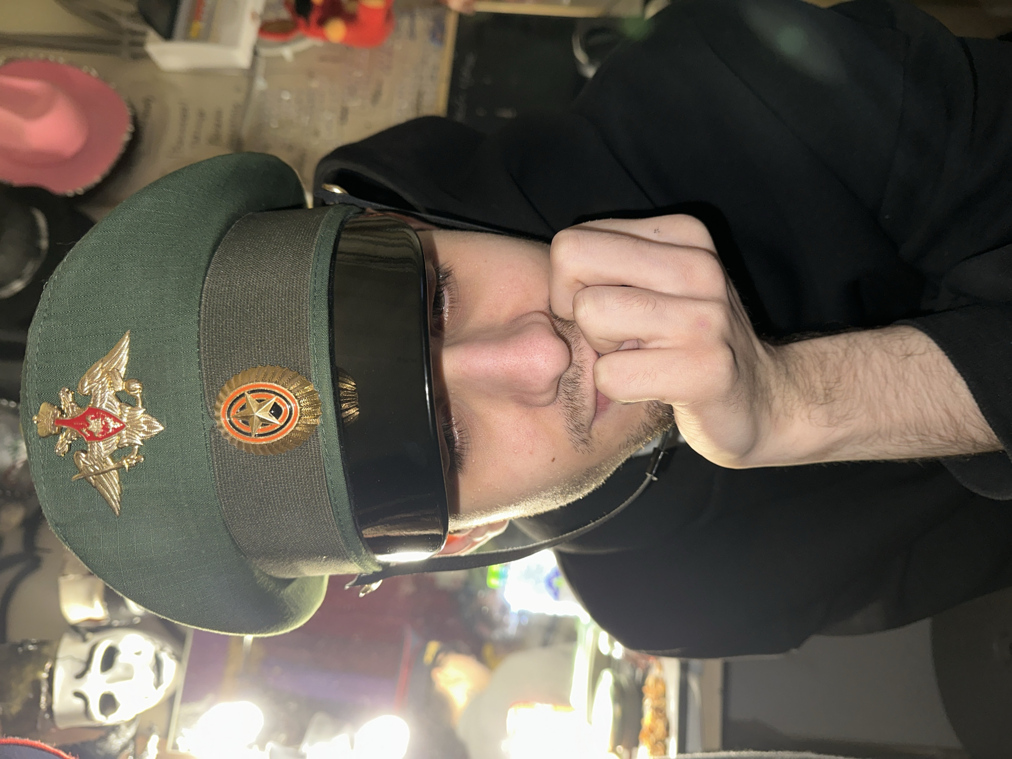 The image features a person, likely male, positioned vertically with their head tilted. They are wearing a dark green military-style peaked cap adorned with two prominent metallic emblems. On the left, there is a large golden double-headed eagle with a shield featuring a red cross and sword. To the right, a circular golden emblem contains a red-outlined star on a dark background. The person is also wearing dark sunglasses that cover their eyes, and a black hoodie or jacket. Their left hand is raised towards their face, with fingers covering part of their mouth and nose, while their gaze is directed towards the viewer. They have light facial hair.
The setting appears to be indoors, with a cluttered background that includes various indistinct objects. Hints in the background show a pink hat on the left, and a white, possibly skull-like mask or decorative item further back on the lower left. The lighting suggests artificial sources, creating reflections on the sunglasses and some bright spots in the background. The overall activity is a posed portrait. The military cap with Russian-style insignia strongly suggests a connection to Russia, which aligns with the given location context of Saratov, Russia. There is no discernible text visible in the image.