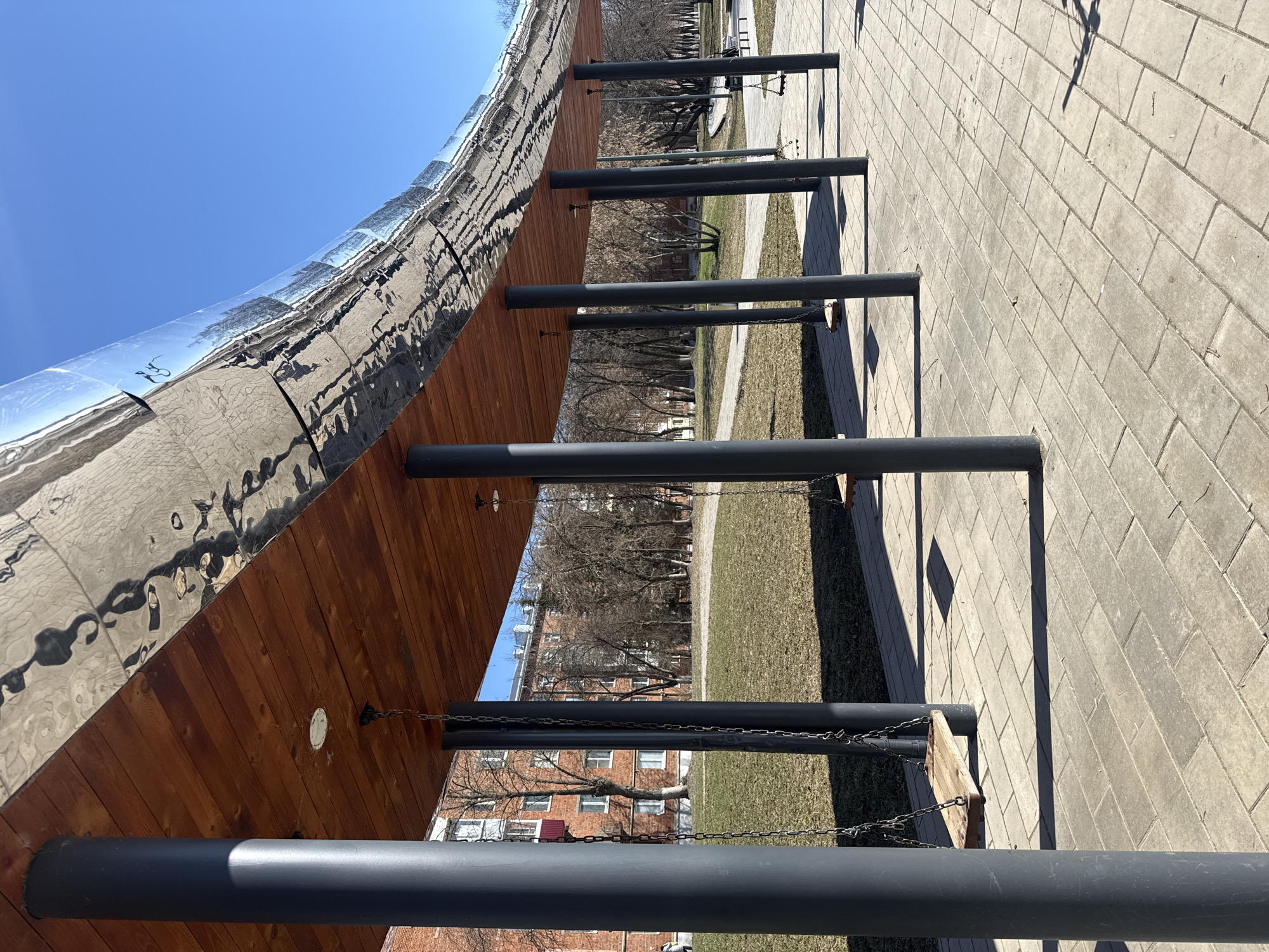 The image depicts a section of a curved structure with a reflective surface, located in what appears to be a park or public space. The structure has a wooden underside, supported by cylindrical, dark-colored posts. Chains are suspended from the wooden structure and attached to wooden boards, possibly swings or seating. The background shows a brick pathway, a grassy area with trees, and multi-story residential buildings. The clear, blue sky indicates it is daytime. The reflection on the structure indicates the presence of people and objects in the environment. The location is in Votkinsk, Russia.