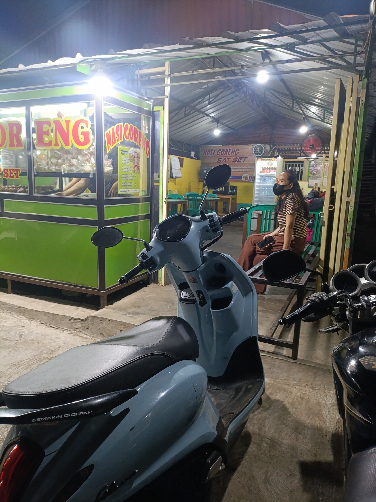 In the city of Jonggol, Indonesia, a low-angle shot captures a nighttime scene outside a food stall. The stall, identifiable by its green and white signage displaying "NASI GORENG" (fried rice) in large red and yellow lettering, is illuminated by overhead lights. Inside, a person is partially visible behind the counter. To the right of the stall, a woman wearing a striped shirt, brown pants, and a black face mask sits on a bench holding a smartphone. She is positioned near a refrigerator stocked with drinks. In the foreground, a light blue scooter is parked, its seat and handlebars clearly visible. Adjacent to it, the front of a black motorcycle is also present. The ground appears to be concrete. The overall environment suggests a casual, open-air dining or takeaway establishment operating in the evening.