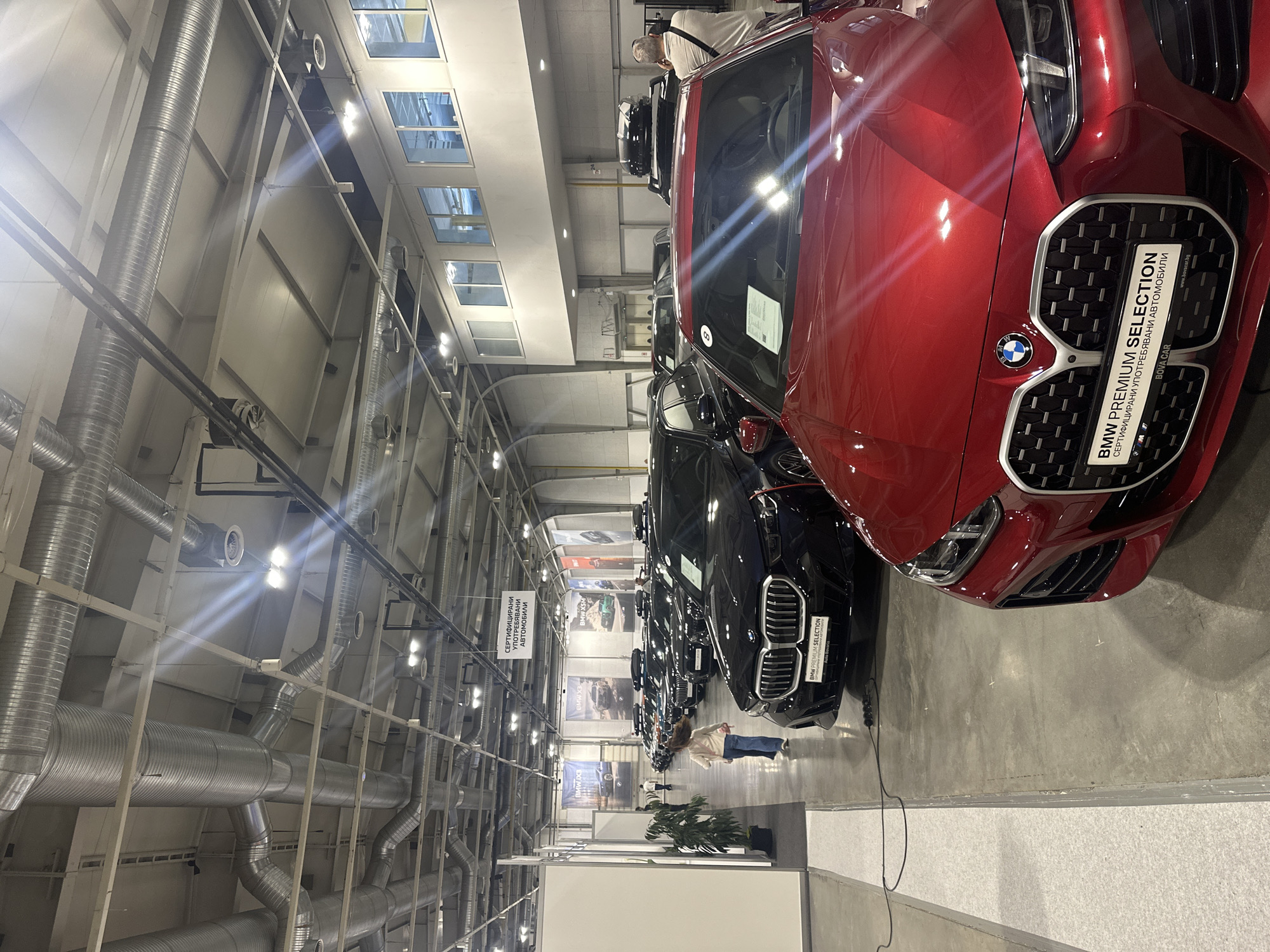An indoor exhibition hall, likely located at the Inter Expo Center in Sofia, Bulgaria, displays multiple BMW vehicles arranged in rows. The scene is illuminated by artificial lighting from numerous fixtures, some of which emit distinct lens flares across the image. The high ceiling features exposed industrial architecture, including extensive silver-colored, corrugated metal ductwork of various diameters, support beams, and a grid of lighting elements.
In the foreground, the front portion of a red BMW G26 4 Series Gran Coupe is visible, angled slightly left, showcasing its large kidney grille with a black mesh pattern and the BMW emblem. A rectangular sign attached to the grille states "BMW PREMIUM SELECTION" in English, and below it, "СЕРТИФИЦИРАНИ УПОТРЕБЯВАНИ АВТОМОБИЛИ" (Certified Used Cars) in Bulgarian, followed by "BOVA CAR www.bovacar.bg". A number "8" sticker is adhered to the windshield.
Behind the red BMW, a dark blue or black BMW SUV (likely an X-series) is prominently featured, facing forward, also displaying a large grille and the BMW emblem. Behind this SUV, multiple other BMW sedans and SUVs, predominantly dark-colored, are parked in rows extending into the background. Several vehicles in the background are equipped with black roof racks or cargo boxes.
A young girl with light brown hair, wearing a white long-sleeved top and blue jeans, walks away from the camera in the mid-ground, moving between a white partition wall on the left and the display cars. To the far right, partially visible, a man in a light-colored shirt and dark trousers stands facing the vehicles, with his back to the camera. Other indistinct figures are visible further in the background among the cars.
On the left side of the hall, a white partition wall creates a boundary. A green potted plant is positioned near this wall. A black electrical cable runs across the light gray concrete floor in the lower left quadrant. Overhead, a rectangular white sign with black Bulgarian text, "СЕРТИФИЦИРАНИ УПОТРЕБЯВАНИ АВТОМОБИЛИ" (Certified Used Cars), hangs above the rows of cars. In the distant background, large banners suspended from the ceiling advertise "BMW X3", "BMW X5", and "BMW X6M" models. The far right wall features a row of elevated windows, through which a light blue ambient glow is visible.