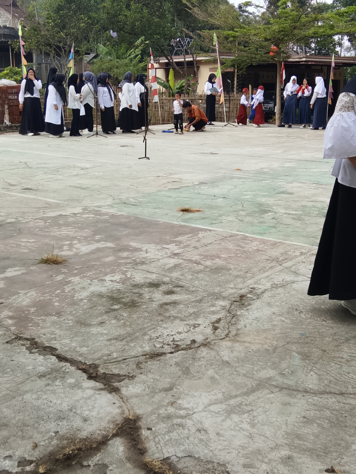 In the city of Jumo, Indonesia, an outdoor scene unfolds on a cracked concrete court, likely an assembly or event ground. A group of students, predominantly girls in white shirts and black hijabs or skirts, stand in formation. To the left, a woman in a black hijab and white top holds a book. Several flags, including a red and white Indonesian flag, are planted in the ground, suggesting a formal gathering. A young child in a white and black checkered shirt is on the court. Another child, in a red cap and skirt, stands near an adult in an orange shirt who is kneeling. In the background, a building with a brown roof and wooden accents is visible, surrounded by trees. The overall atmosphere suggests an event or ceremony taking place.