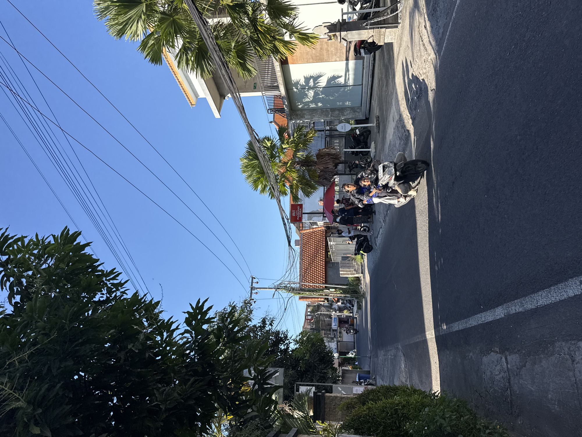The image shows a street in North Kuta, Indonesia, lined with buildings and vegetation. Motorbikes are parked on the side of the road. Overhead, power lines are visible against a clear blue sky. Palm trees and other greenery are scattered along the street. A person sits on one of the motorbikes. The road is paved and has a white line marking. Buildings vary in style, some with red tile roofs and others with more modern facades.