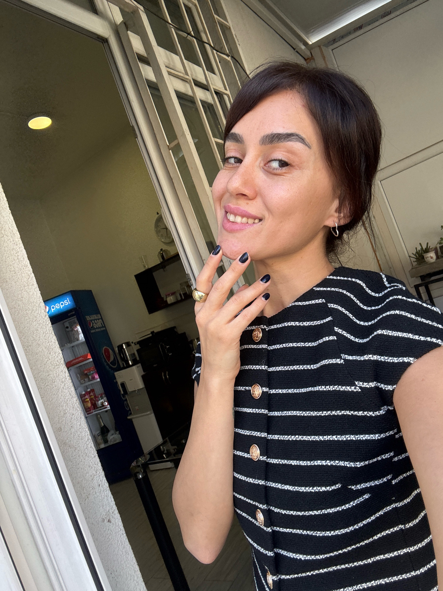 A woman is taking a selfie in what appears to be a residential or commercial building, possibly in Tbilisi, Georgia, judging by the context. She has dark hair and is wearing a black and white striped top with gold buttons. She is smiling and touching her face with her left hand, which has dark nail polish and a gold ring. She is also wearing a small silver hoop earring. In the background, through an open doorway or window, a Pepsi branded refrigerator is visible, stocked with beverages and food items. Beyond the refrigerator, there are shelves with various items, a clock on the wall, and what looks like kitchen appliances. The lighting suggests it is daytime.