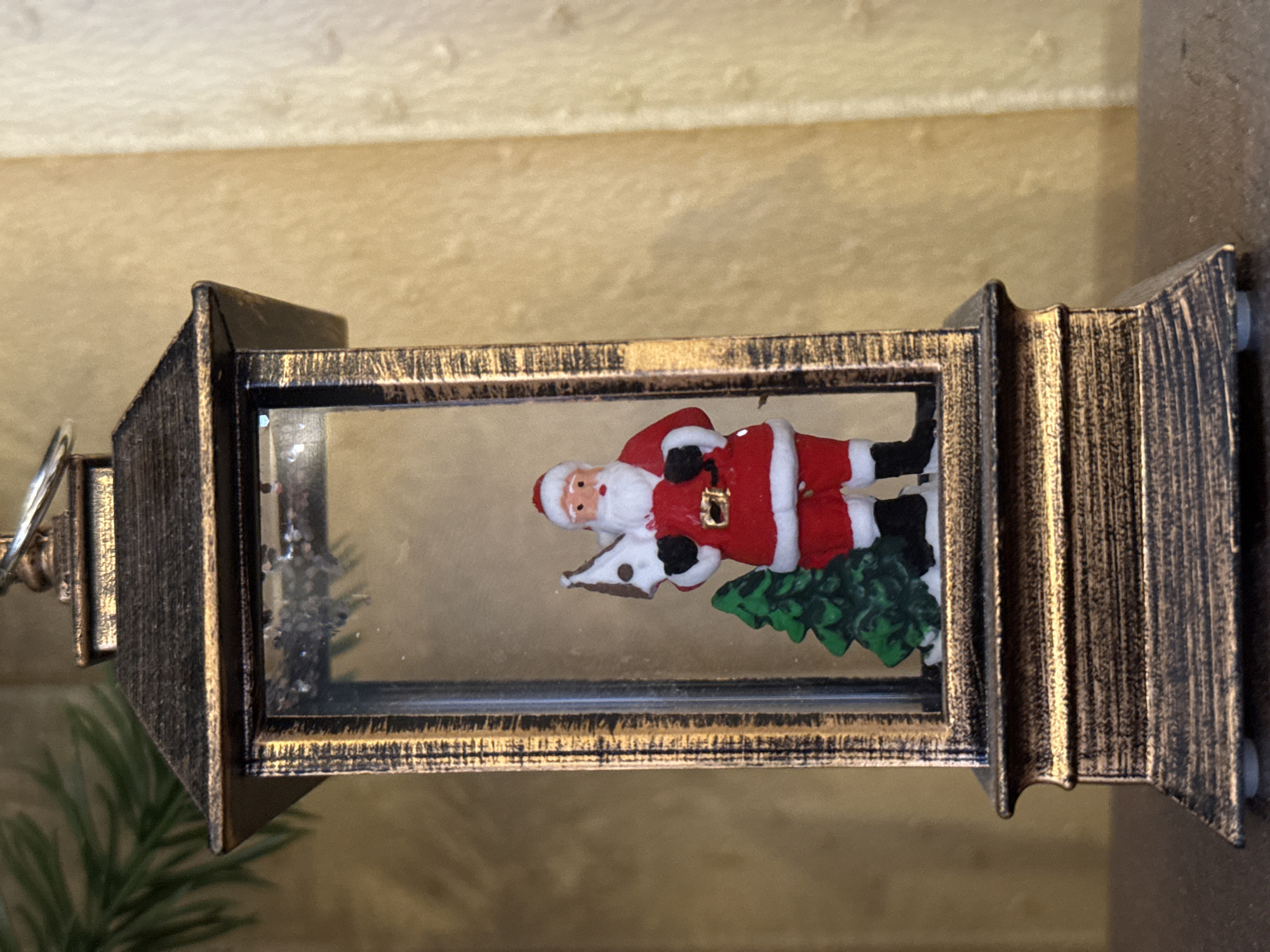 An outdoor Christmas decoration featuring a miniature Santa Claus holding a Christmas tree and standing next to a larger Christmas tree inside a metallic lantern. The lantern has a dark, antique finish with gold accents. The background is a blurred golden textured wall, and a blurred green pine branch is visible in the upper left corner. The lantern sits on a dark surface. The image was taken in Khimki, Russia.