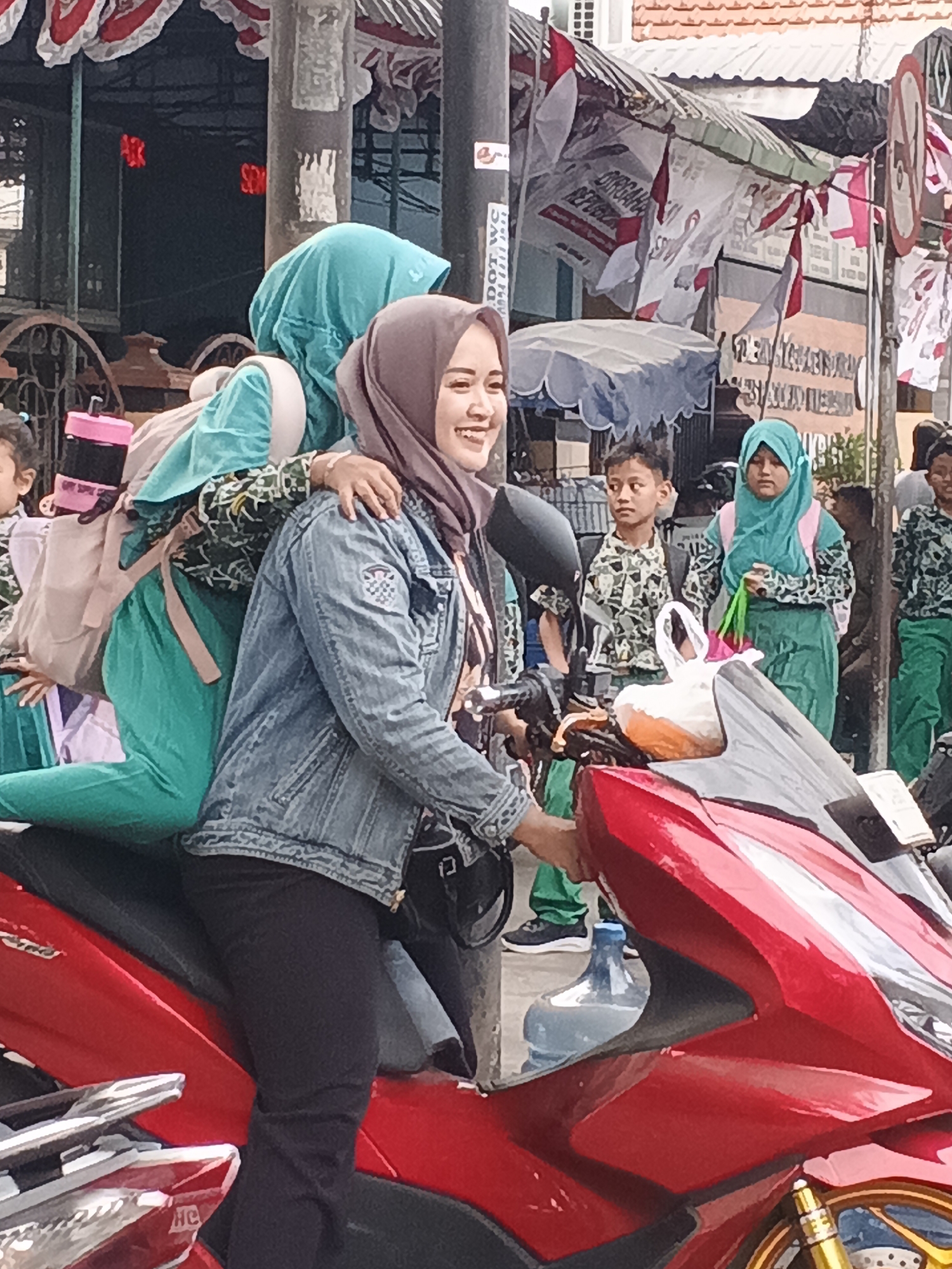 In the city of Krian, Indonesia, on a street, two young women wearing hijabs are seen on a red motorcycle. The woman in the foreground, wearing a denim jacket and black pants, has a grey hijab and is smiling while looking to her right. She has her right arm around the other woman. The second woman, with a teal hijab and a patterned green and white shirt, is seated behind her, also wearing green pants and carrying a pink backpack. The background reveals a bustling street scene with other individuals, some appearing to be students in school uniforms, and banners with Indonesian flags and text. The environment suggests a public gathering or a normal day on a busy Indonesian street.