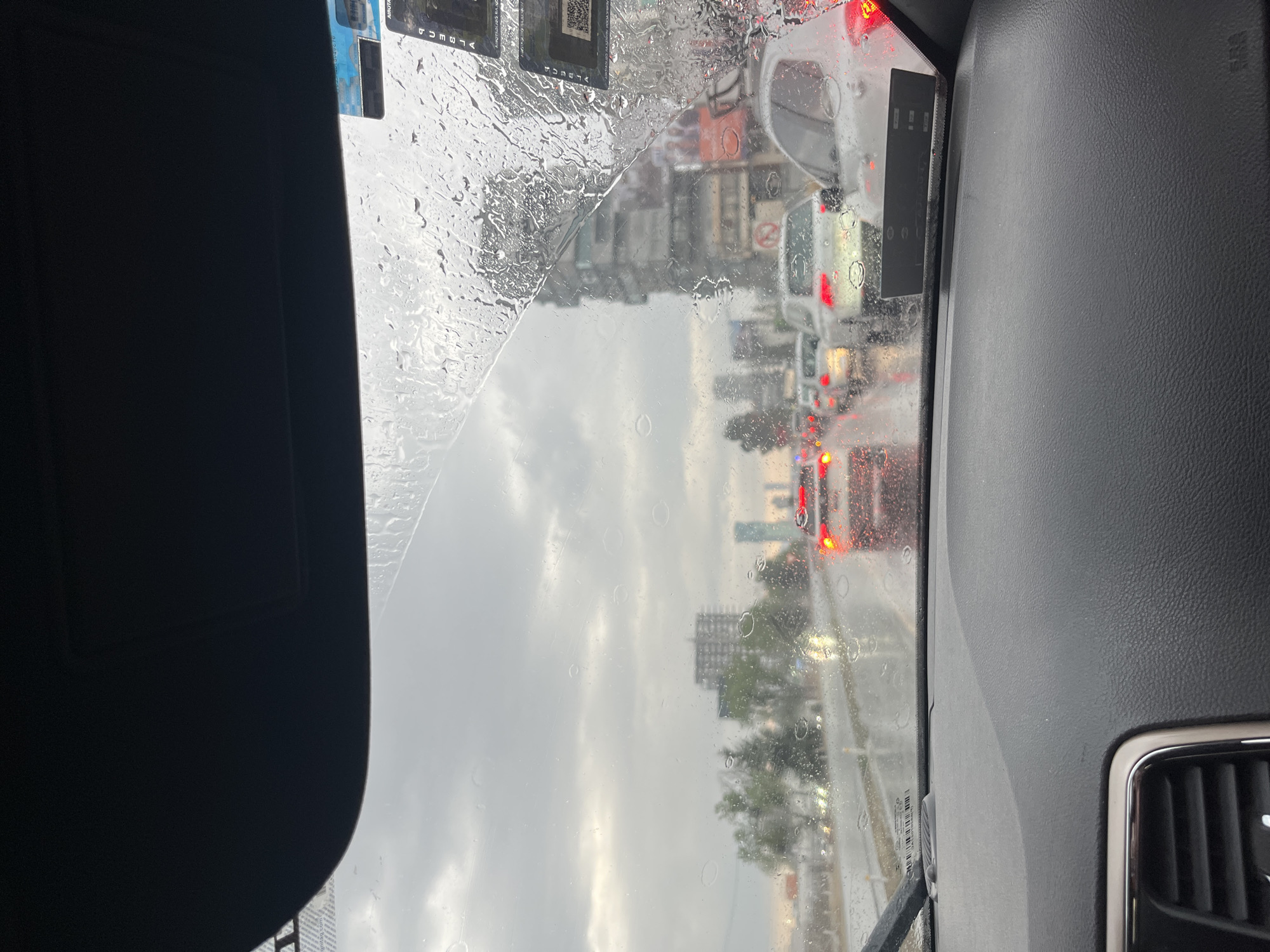 The image, taken from inside a vehicle, shows a rain-streaked windshield with traffic and city buildings visible through the blurred view. Raindrops heavily cover the glass, distorting the urban landscape. In the foreground, the interior of the car is visible, including parts of the dashboard, sun visor, and interior components. The scene depicts heavy traffic on a rainy day in an urban setting. The buildings in the background appear to be in San Bernardino Tlaxcalancingo, Mexico. The buildings seem to be of medium height. In the middle distance, traffic with visible brake lights suggests congestion. The windshield wiper is partially visible, indicating its use.