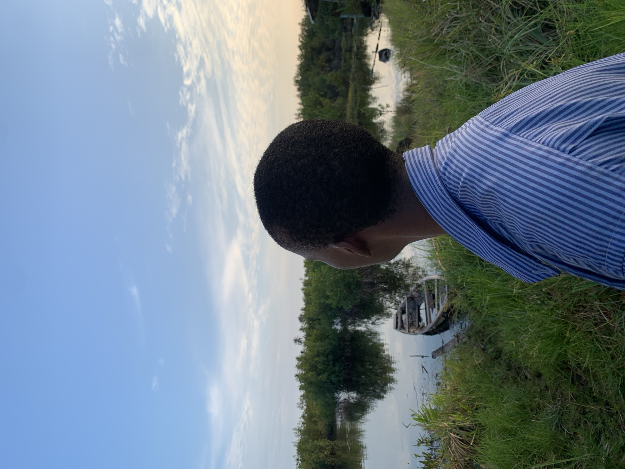An outdoor scene captures a person observing a natural landscape, likely a river or lagoon, under a late afternoon or early evening sky. The image is framed from behind the individual, showcasing the back of their head and right shoulder. The person, appearing to be male, has short, dark, curly hair and is wearing a blue and white striped collared shirt.
They stand on a lush, green bank, looking out over a calm body of water. The water reflects the soft colors of the sky, which transitions from a clear blue on the left to warmer, golden hues with scattered white clouds towards the right, hinting at an approaching sunset.
On the far bank, dense green trees and mangroves line the water's edge. An old, weathered wooden boat, appearing somewhat derelict, is partially visible in the water near the bank, adding a rustic element to the scene. The overall atmosphere is tranquil and contemplative. There is no visible text in the image. The setting is consistent with a natural waterway in a tropical region, such as Ibasa, Nigeria.