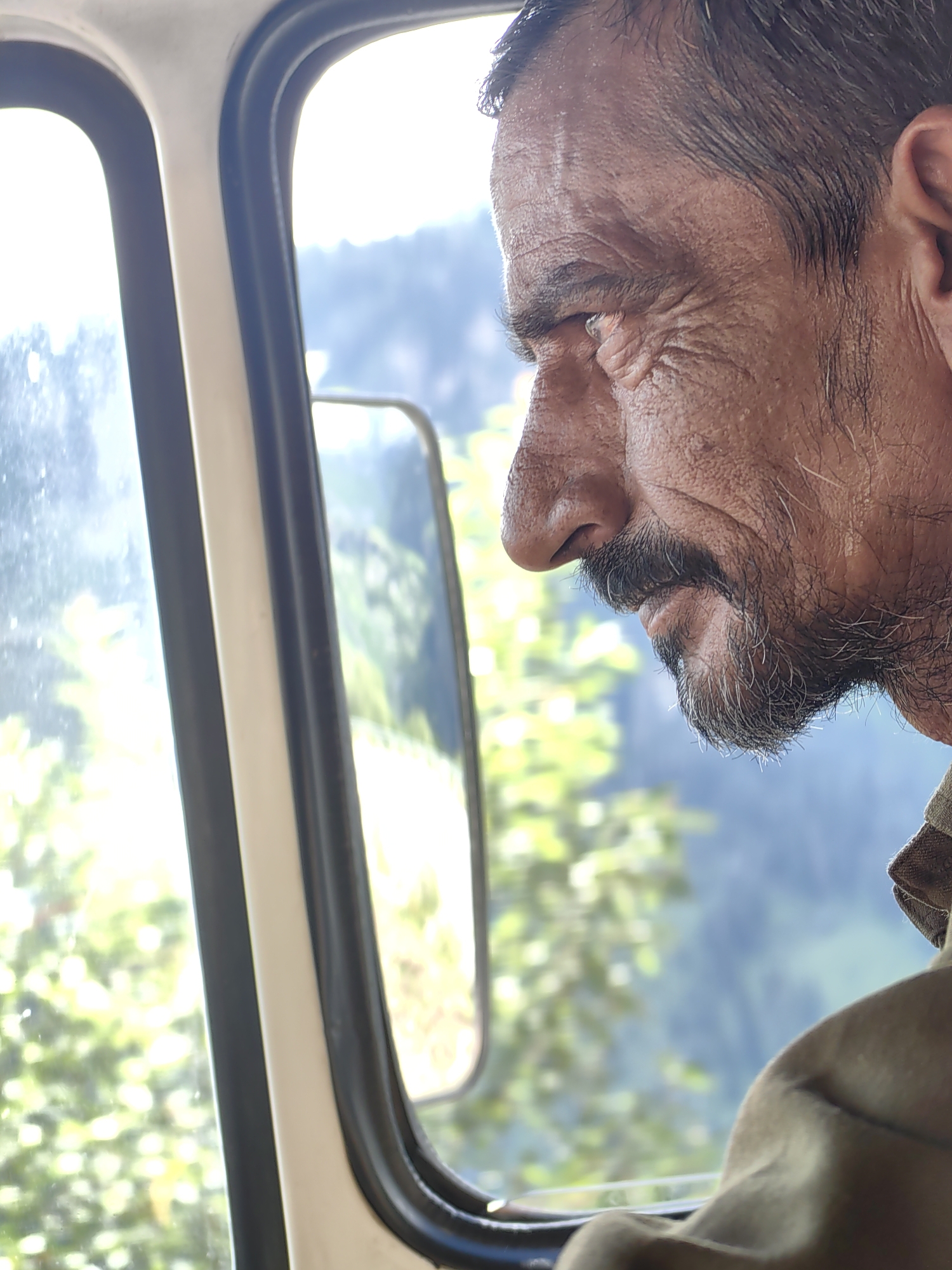 A medium shot shows a man's weathered face in profile, looking out a vehicle window. The man, who appears to be of Indian descent and is likely in his 50s or 60s, has deep wrinkles around his eyes and forehead, a prominent nose, and a salt-and-pepper beard and mustache. He is wearing a brown collared shirt. The window frames his face, with the left side of the image showing a blurred view of green foliage and a bright sky, suggesting an outdoor setting in a mountainous region. A side mirror is visible in the background, reflecting more of the landscape. The image is taken from inside a vehicle, possibly a truck or van, as indicated by the window frame and interior paneling. The man's gaze is directed outwards, implying he is a driver or passenger observing the surroundings. The scene is likely set in Kothi, India, a location known for its scenic beauty and mountainous terrain.