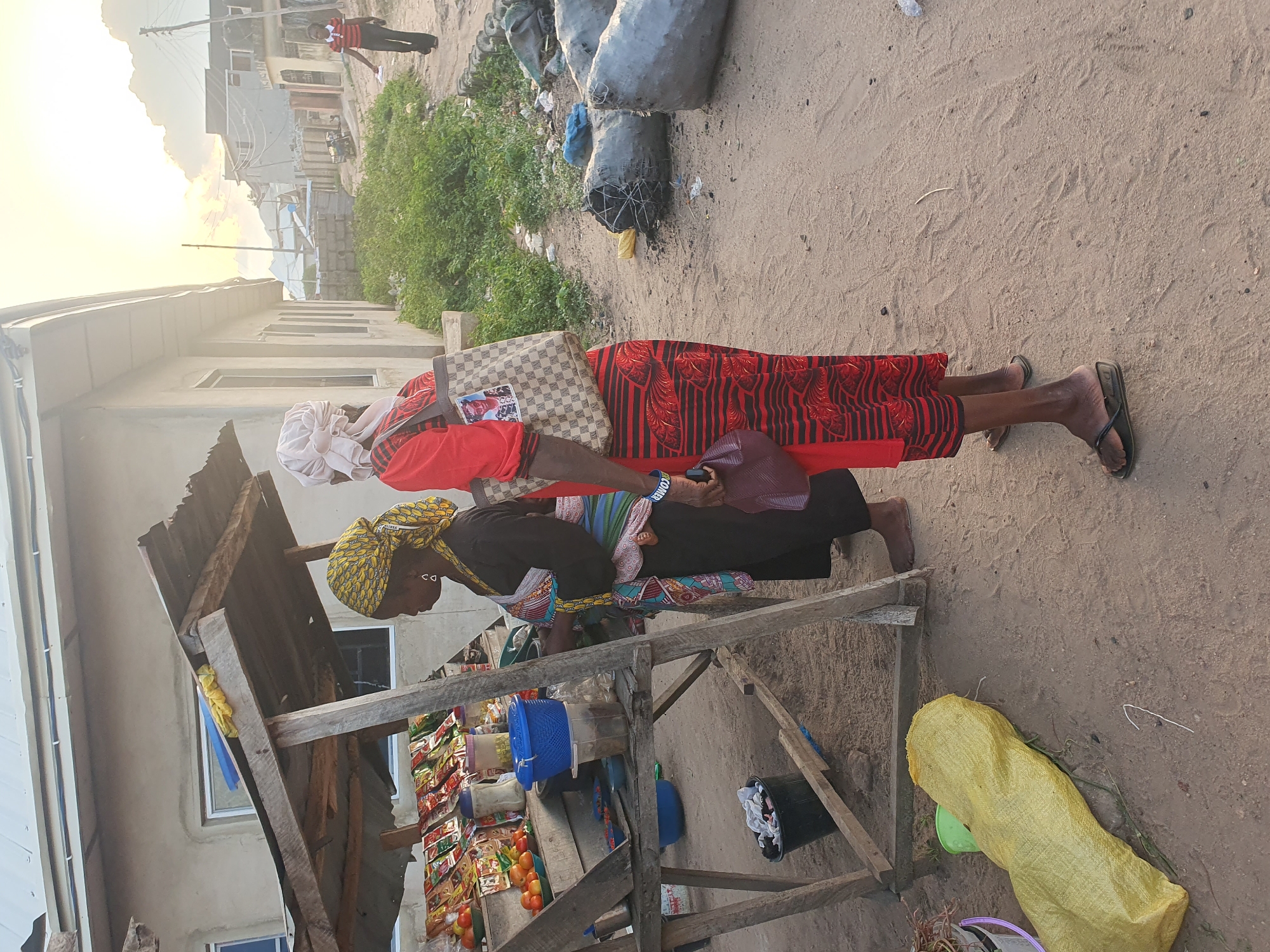 The image depicts an outdoor scene in Lokoja, Nigeria, at dusk. Two women are standing near a makeshift market stall constructed from wood and corrugated metal. The stall displays various packaged goods and some fresh produce, including tomatoes. One woman, on the left, wears a black dress with a colorful patterned wrap and a yellow headscarf. She has earrings and appears to be interacting with the stall. The second woman, on the right, is wearing a red dress with black stripes and a white headscarf. She is carrying a large patterned tote bag and a dark-colored handbag.
The ground is sandy and appears to be a dirt road. In the background, a building with a light-colored facade and windows is visible, along with other structures and vegetation. Several large, dark, cylindrical objects, possibly sacks or drums, are lying on the ground to the right of the women. A yellow sack is also present on the ground to the left of the stall. A young boy in a red and white striped shirt and dark pants walks away in the background towards the right. The sky is illuminated by the setting sun, casting a warm glow on the scene.