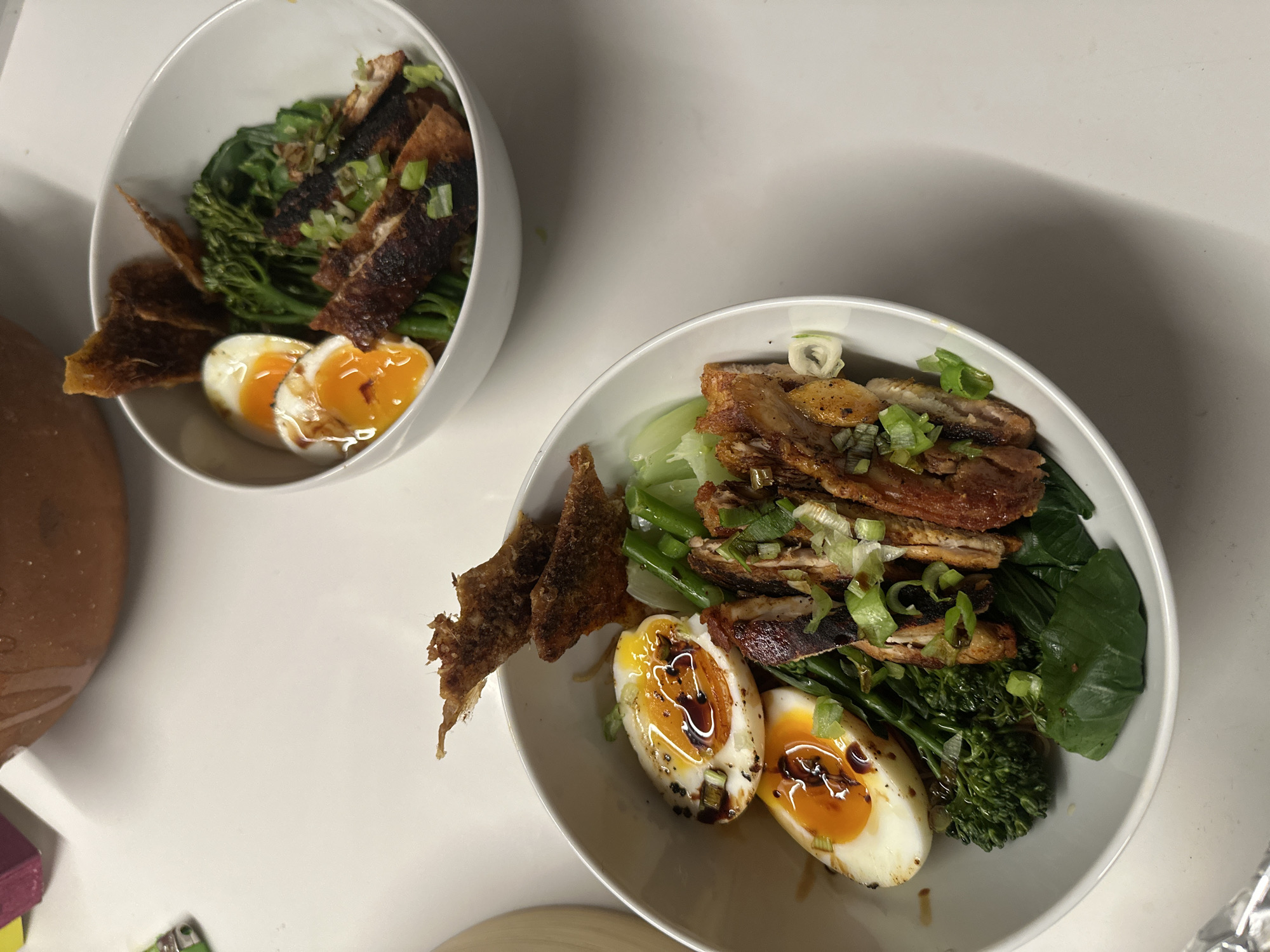 Two white bowls containing meals are positioned on a white surface. Each bowl appears to contain sliced meat, eggs, green vegetables, and crispy fried elements. The yolks of the eggs are runny and appear to have been drizzled with a dark sauce. Chopped green onions are scattered over the meals. The general composition suggests a healthy, balanced meal, possibly a variation of ramen or a similar Asian-inspired dish. Other objects like a brown pot and other cooking-related items are visible in the background, partially out of focus. No people are visible. The environment appears to be a kitchen or dining area. There is no indication as to the location in London, United Kingdom.