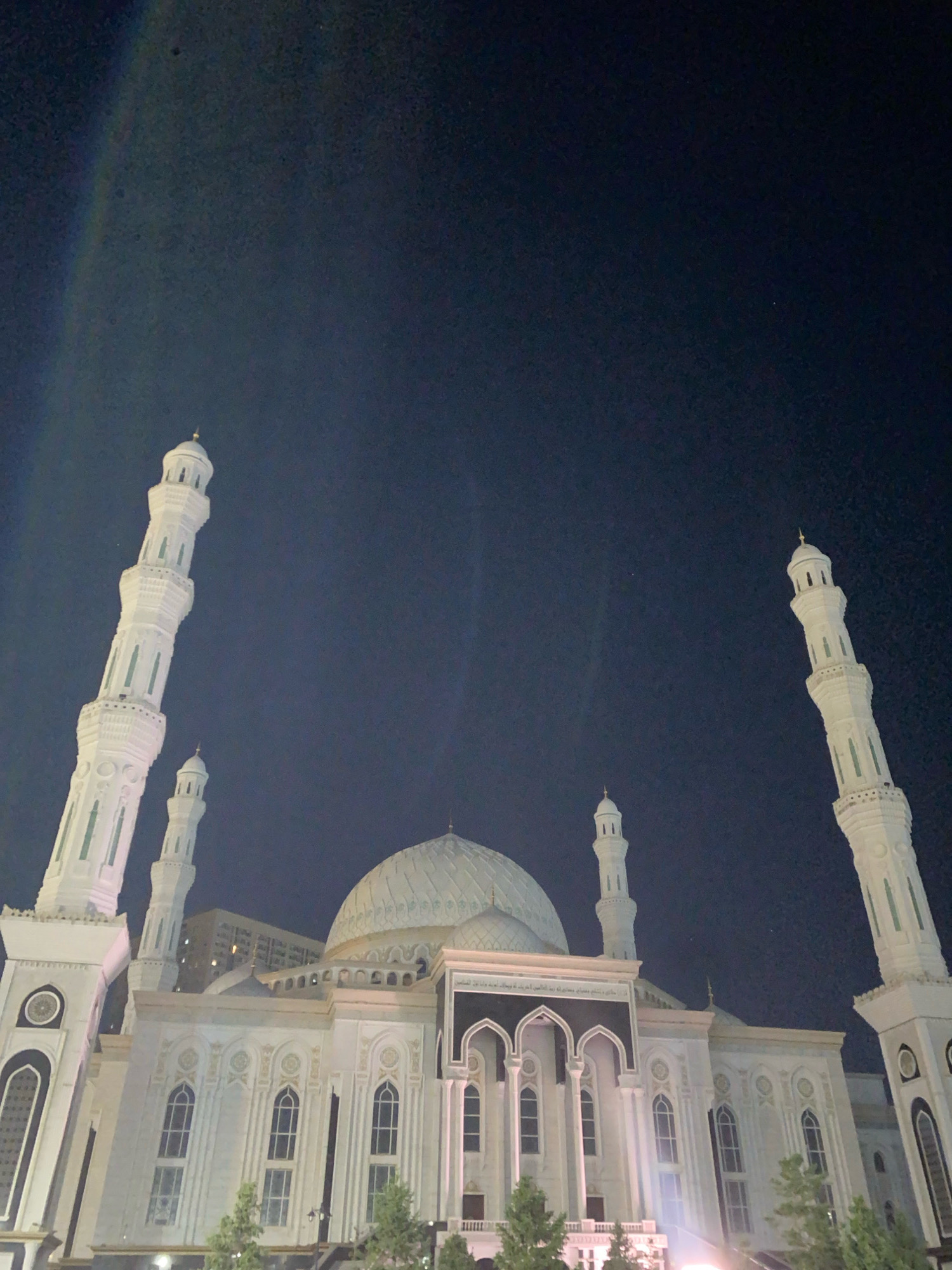 An exterior, low-angle, night-time, wide shot depicts the Hazret Sultan Mosque in Astana, Kazakhstan. The mosque is illuminated by artificial lights, highlighting its white facade, intricate carvings, and prominent dome. Two tall minarets, also illuminated, flank the main structure. The night sky is dark, with faint streaks of light visible in the upper left corner, possibly from streetlights or light pollution. The mosque's architectural details, including its numerous arched windows and decorative elements, are clearly visible against the dark background. In the foreground, the tops of trees can be seen. The image is slightly blurred, suggesting it may have been taken with a handheld camera or under low-light conditions.