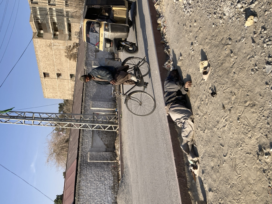 The image depicts an outdoor urban scene in Quetta City Tehsil, Pakistan, under a clear blue sky. In the immediate foreground, an adult male is lying prone on a dirt shoulder adjacent to a paved road. He is dressed in light-colored trousers and a dark long-sleeved garment, with his head turned away or pressed into the ground. Two light-colored sandals, a small dark object, and a brick fragment are visible on the dirt near him.
On the paved road, an adult male with a beard and cap is seated on a black bicycle, appearing to be stationary or moving slowly. He wears a dark blue jacket and light brown shalwar, and a white sack is secured to the bicycle's rear rack. His gaze is directed forward and slightly to his right. To the right and slightly behind the bicyclist, a yellow and black three-wheeled motorized rickshaw is partially visible on the road, with its driver discernible inside.
In the mid-background, a low white-painted brick wall with a corrugated metal roof runs parallel to the road. A metal lattice tower with a green flag attached near its top stands close to the wall. Further in the background, a multi-story building constructed from light-colored stone or concrete is visible under the clear sky. Bare trees are also present in the background.