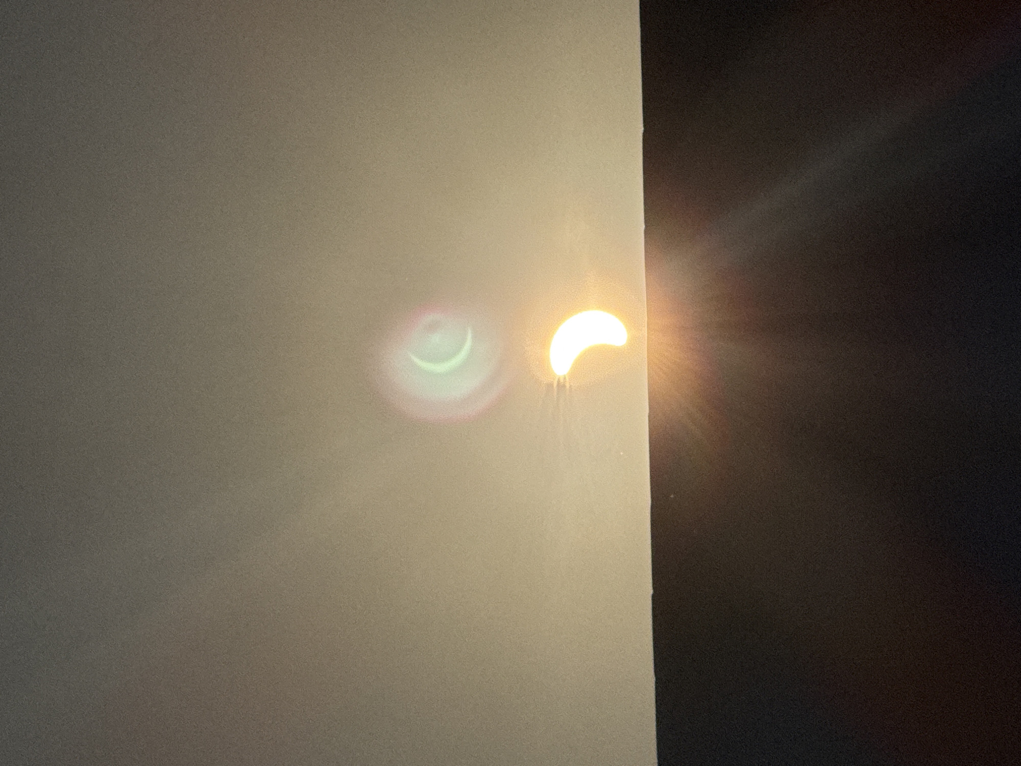 The image depicts a partial solar eclipse occurring over the town of Great Yarmouth, United Kingdom. The sun, partially obscured by the moon, appears as a bright crescent shape near the horizon. Above the sun, a faint, crescent-shaped reflection is visible, possibly due to atmospheric conditions or the camera lens. The foreground is dominated by a dark, horizontal surface, likely a building's roof or a wall, which casts a shadow. The sky is hazy with a gradient of light from the horizon upwards. No individuals or specific landmarks of Great Yarmouth are identifiable in this image.