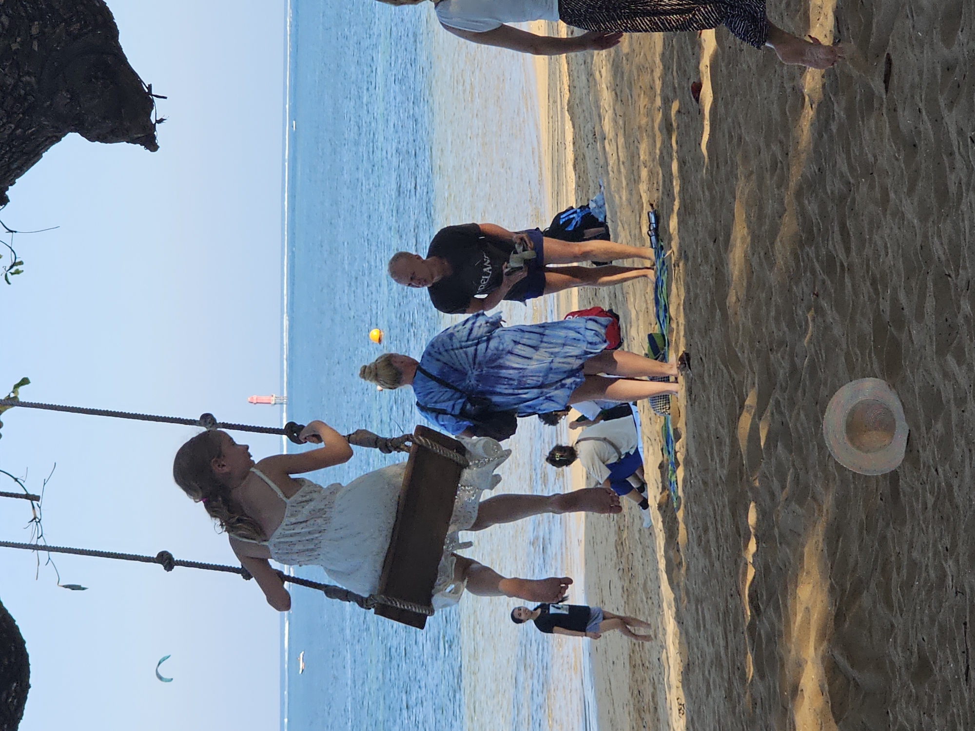This media file captures a family enjoying a day at the beach. The scene is set on a sandy beach with the ocean in the background. The sun is high in the sky, indicating it's likely daytime.
In the foreground, a young girl is joyfully swinging on a rope swing suspended from a tree. She is wearing a light-colored dress and appears to be mid-swing, with her legs outstretched. Her face is turned to the side, and she seems to be smiling.
Further back, several other individuals are visible. A man in a black t-shirt and blue shorts is holding something in his hands, looking down. Another person, wearing a blue tie-dye shirt, stands near him. A person in a white t-shirt and blue shorts is also present, along with a child in a black t-shirt and shorts. The people are gathered on the sand, some appearing to be walking or standing. A straw hat rests on the sand to the right of the frame.
In the distance, the calm blue ocean stretches out to the horizon, with a faint line of white where waves are breaking. A few kite surfers are visible on the water, adding a touch of activity to the otherwise tranquil scene. The overall mood appears to be relaxed and happy, with people enjoying a leisurely day at the beach. There is no visible text in the image. The location is Denpasar, Indonesia.