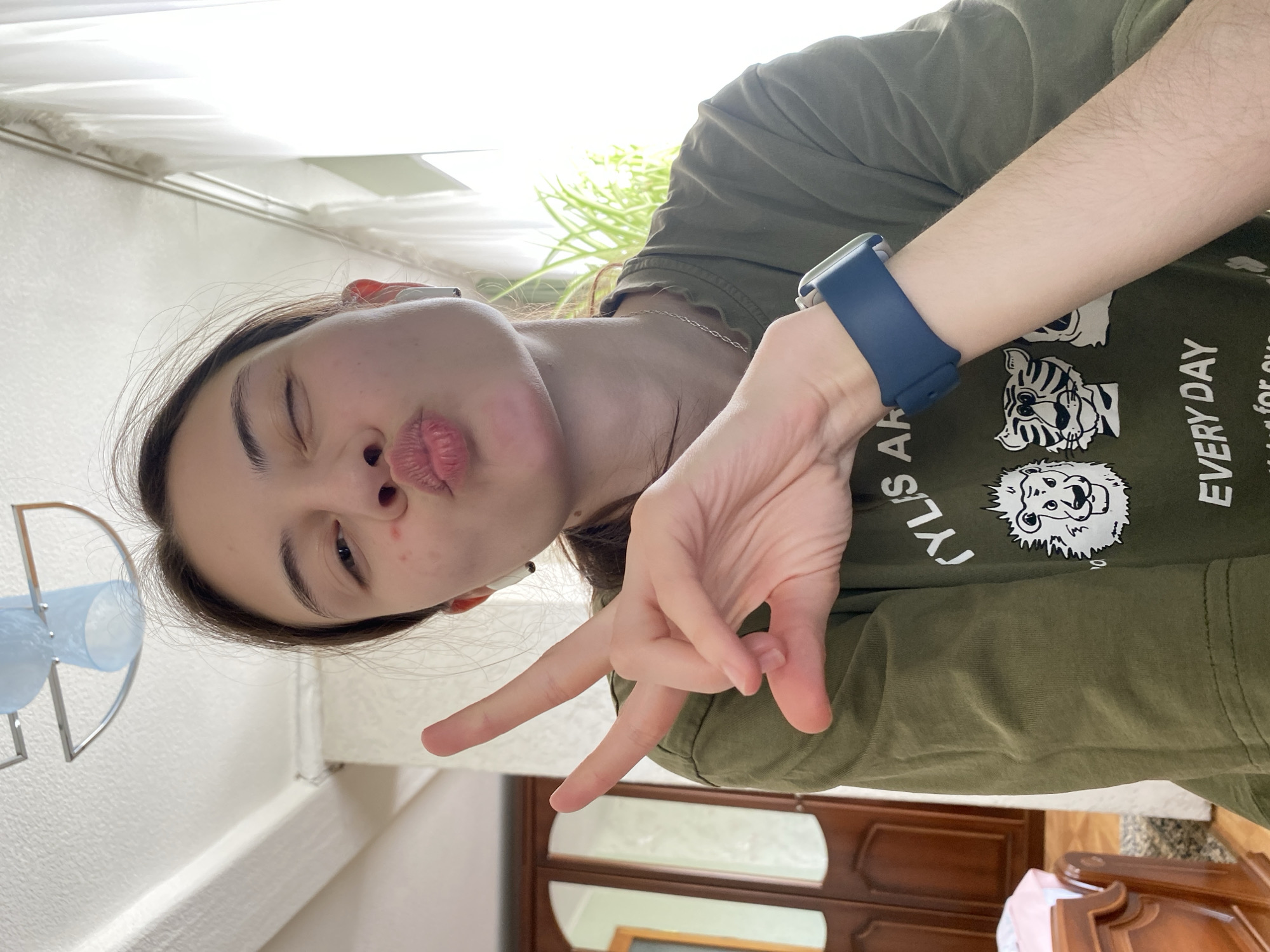 A young woman is posing for a selfie indoors. She is wearing a green t-shirt with cartoon animal prints and the word "EVERYDAY" visible. She is making a kissy face and holding up a peace sign with her right hand, which also has a blue smartwatch on her wrist. She has fair skin, dark hair, and is wearing what appears to be wireless earbuds. The background includes a window with daylight shining through, indoor plants, and a partial view of dark wooden furniture. The room appears to be a bathroom, with a metal and plastic bathroom organizer visible. The location is indeterminate and there is no evidence it is in Votkinsk, Russia.