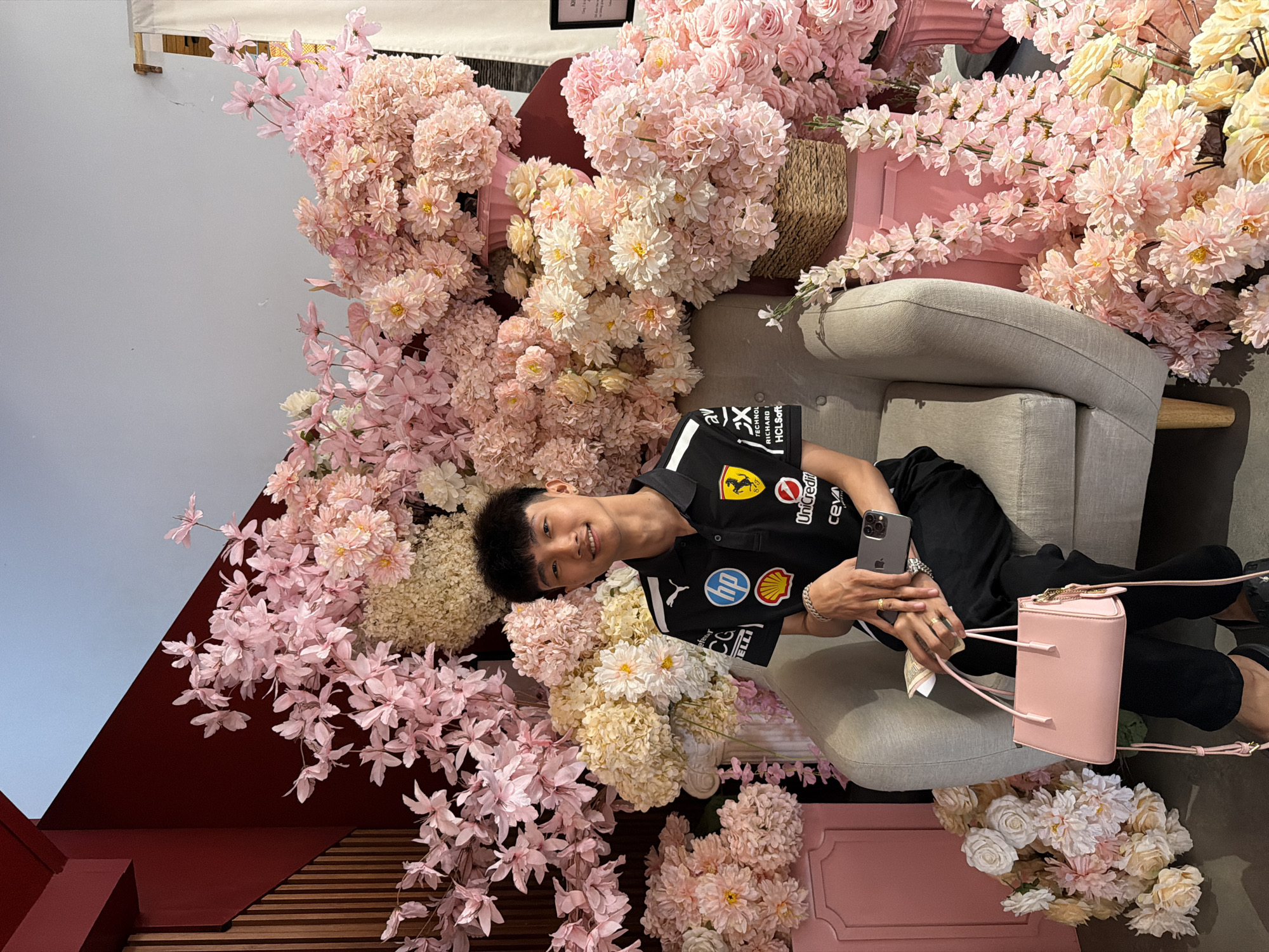 An East Asian man is depicted in an indoor setting, reclining comfortably on a light-colored upholstered armchair, surrounded by an extensive arrangement of artificial flowers. He has short black hair and is smiling warmly at the camera. He is dressed in a black polo shirt featuring numerous sponsor logos, including Ferrari (with its emblem), UniCredit, Puma, HP, Shell, Ceva, HCLSoft, and 2X Technology, paired with black trousers.
The scene is dominated by dense, decorative clusters of artificial flowers in various shades of pink, light pink, cream, and white, resembling cherry blossoms, chrysanthemums, and roses. These abundant floral displays are arranged around and behind the armchair, creating a lush, vibrant, and soft backdrop. Elements of a dark red wall or partition and a light blue-grey wall are also visible in the background, complementing the floral decor.
The man holds a silver smartphone in his left hand. A small, structured pink shoulder bag with gold hardware rests on the sofa near his right hand. He is also wearing rings on his right hand and a bracelet on his left wrist. The overall impression is a posed photograph in a specially designed photo opportunity area, suggesting a festive or aesthetically pleasing commercial space within Quy Nhon Ward, Vietnam. The time of day appears to be daytime, with a cheerful and inviting atmosphere.