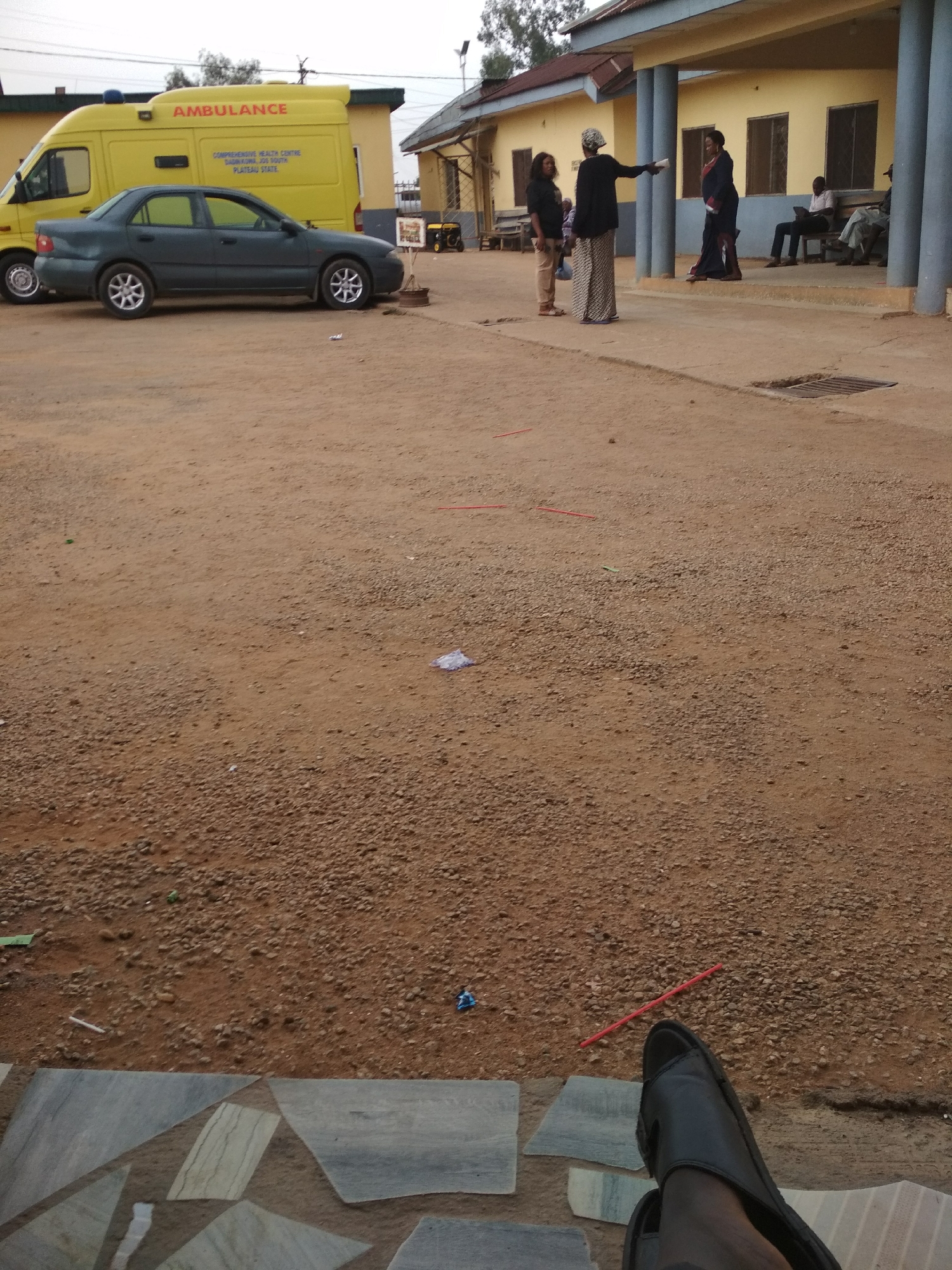 The scene shows the exterior of the Comprehensive Health Centre in Langtang South, Plateau State, Nigeria. An ambulance and a dark green car are parked in front of a one-story building with yellow walls and a brown roof. Four women and two men are standing and sitting near the building's entrance. One of the women is gesturing with a piece of paper. The ground is a reddish-brown color with loose gravel. Several pieces of trash are scattered around. In the foreground, a person is wearing a black sandal. The background shows trees and the sky.