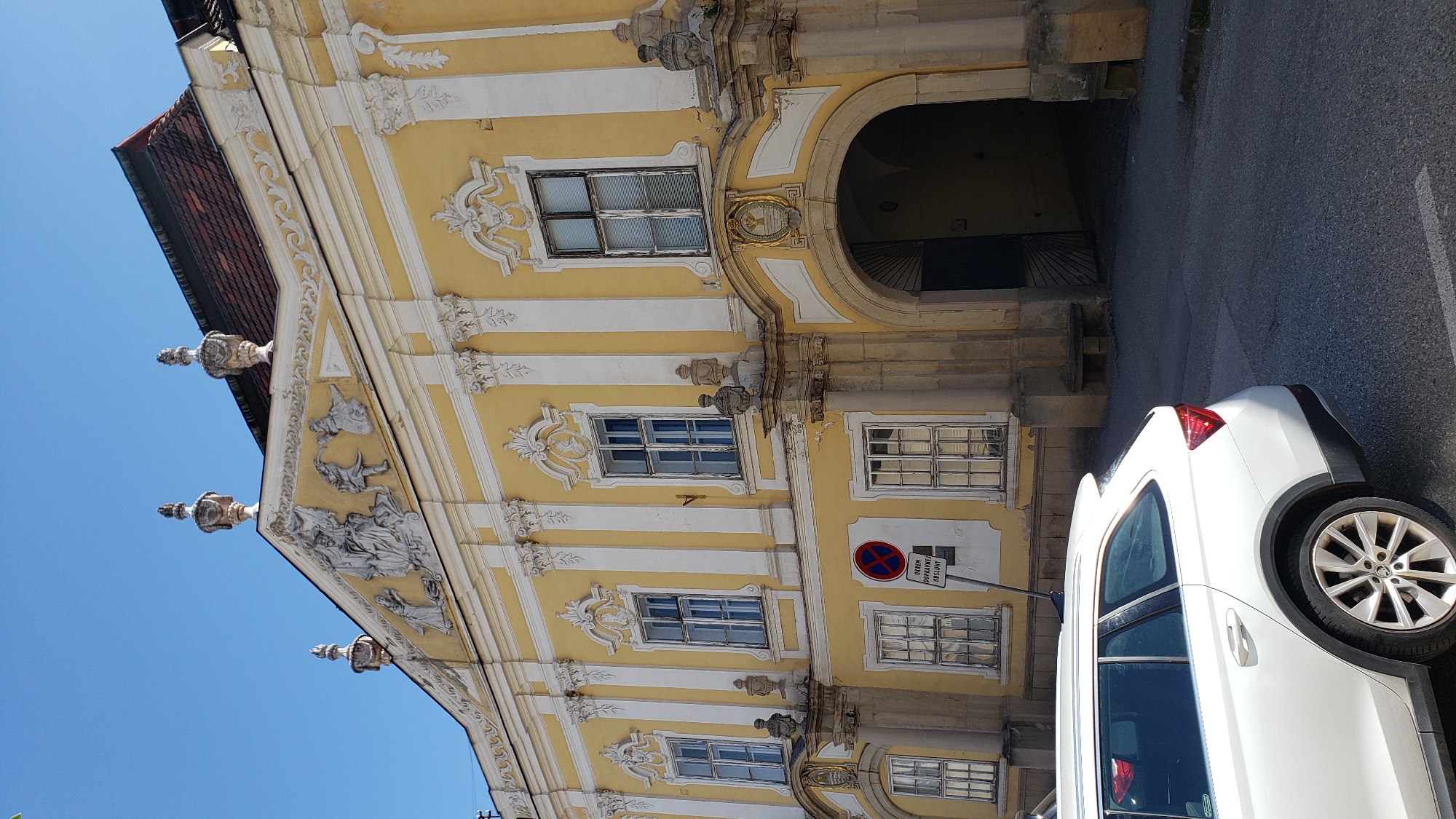 The image depicts the exterior of the Rákóczi family mansion in Prešov, Slovakia. The facade is painted yellow with white decorative elements and features multiple windows. A white car is parked in the foreground on the asphalt road. A traffic sign indicating "No stopping, except for delivery service" is visible to the left of the building's arched entrance. The architecture suggests a historical Baroque or Neoclassical style. The sky is clear and blue.