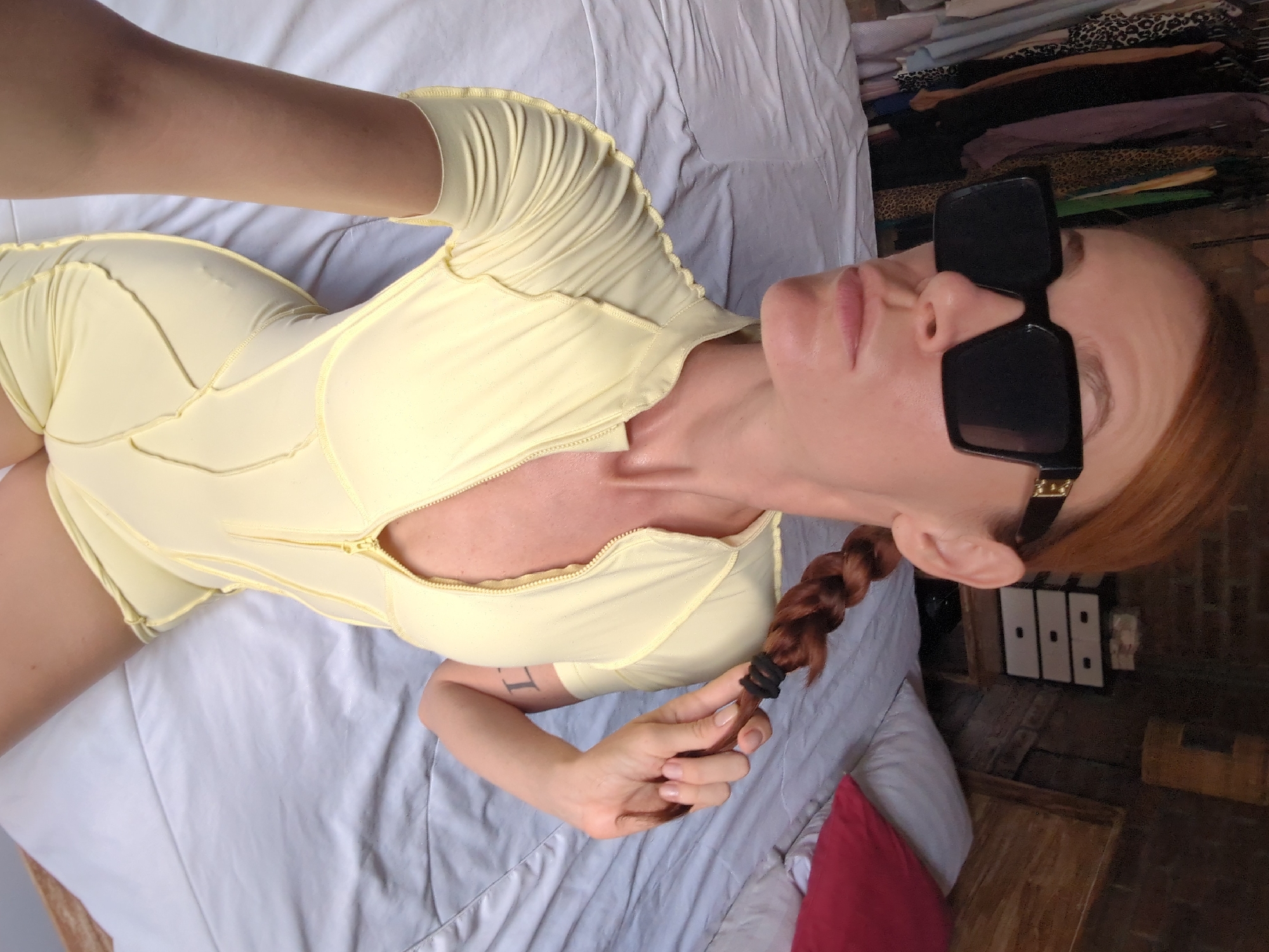 A woman with red hair braided into a single plait lies on a bed covered in white sheets. She wears large black sunglasses and a form-fitting, short-sleeved, pale yellow playsuit with a zip-up front. Her left arm is visible, and she is holding the end of her braid in her left hand. Part of a red pillow is visible on the bed. In the background, a wooden floor, a chest of drawers, and folded clothing are visible. The photograph was taken in North Kuta, Indonesia.