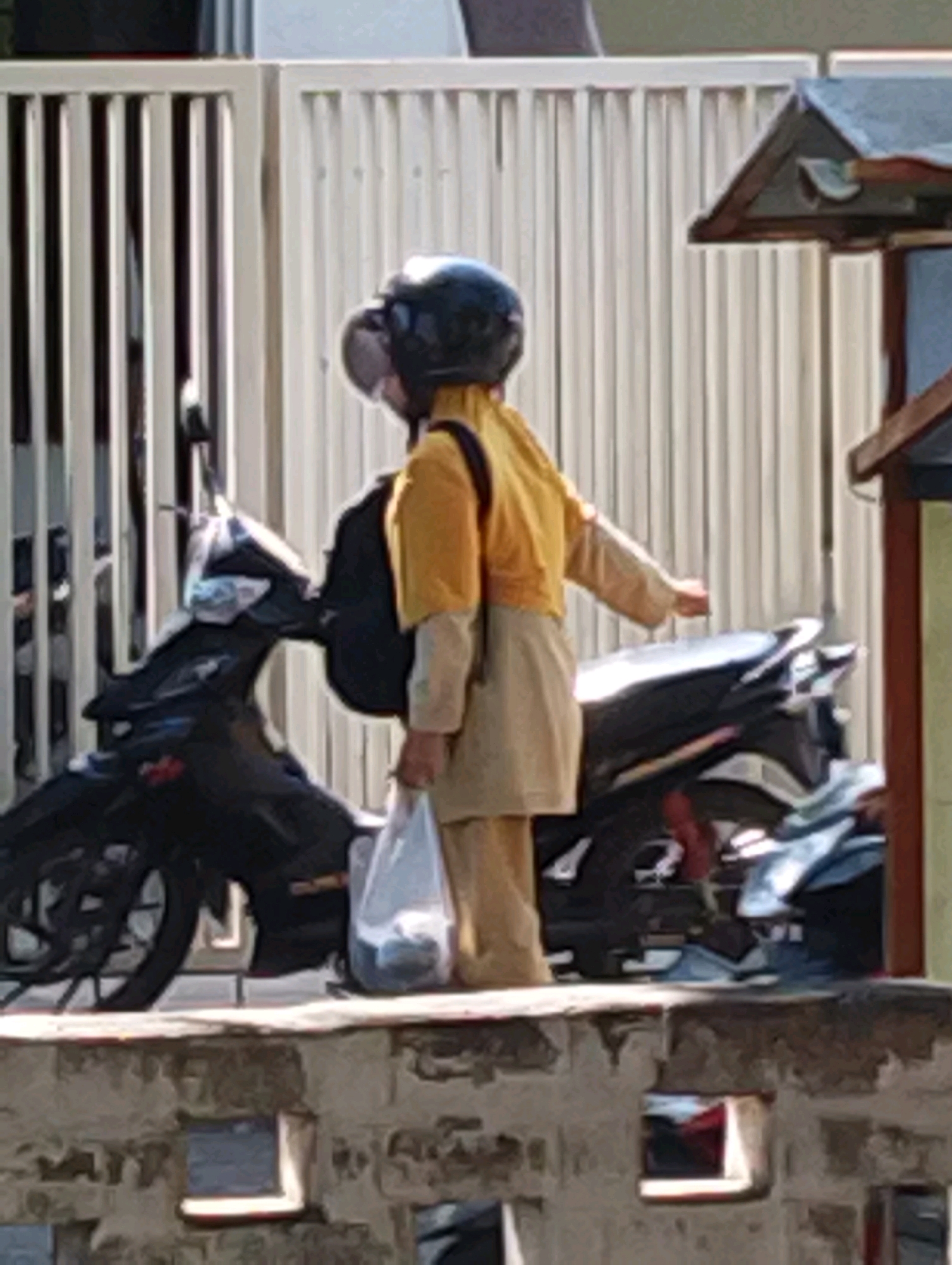 In Bangil, Indonesia, a person wearing a yellow jacket and a dark helmet stands next to a black motorcycle. The person is holding a white plastic bag in their right hand and has a black backpack on their back. The motorcycle is parked in front of a white fence, and there is a partial view of a building with a dark roof on the right side of the image. The ground appears to be paved.