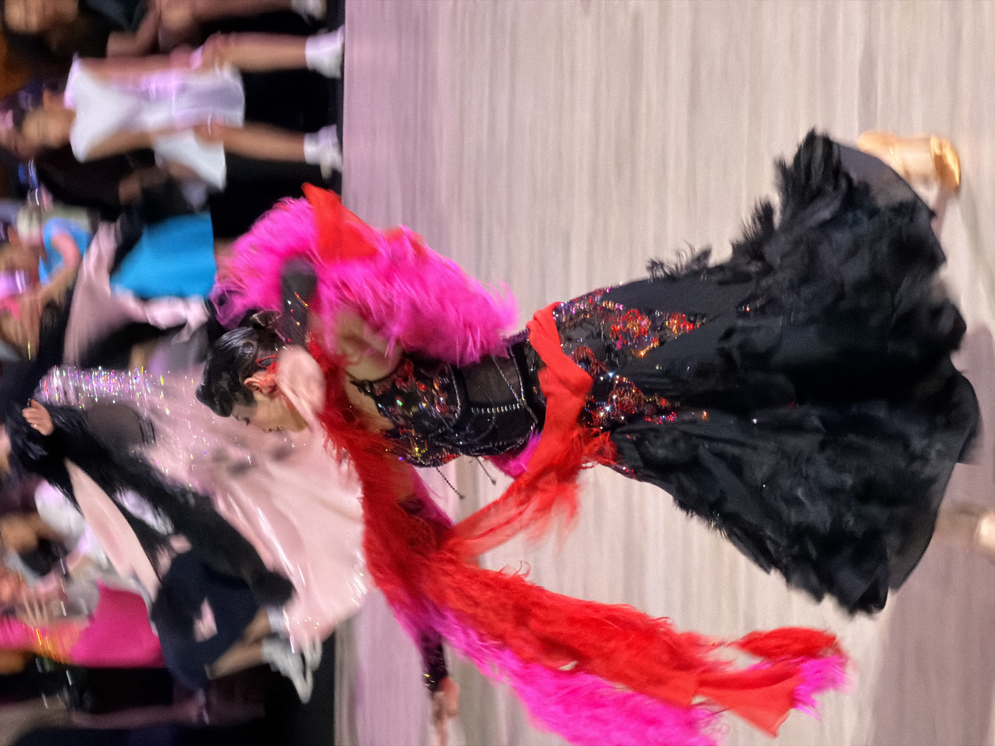 The image shows a dancer in a black and red feathered dress with a red and pink feathery boa, performing on a wooden floor. The dancer is wearing a black corset with red and gold embellishments and has dark hair pulled back. In the background, other people are visible, some in brightly colored dresses and others in white. The scene appears to be a ballroom dancing competition or performance, possibly in a large hall with a polished floor. The lighting is bright, suggesting an indoor venue. There is no indication that the location is in Hà Nội, Vietnam.