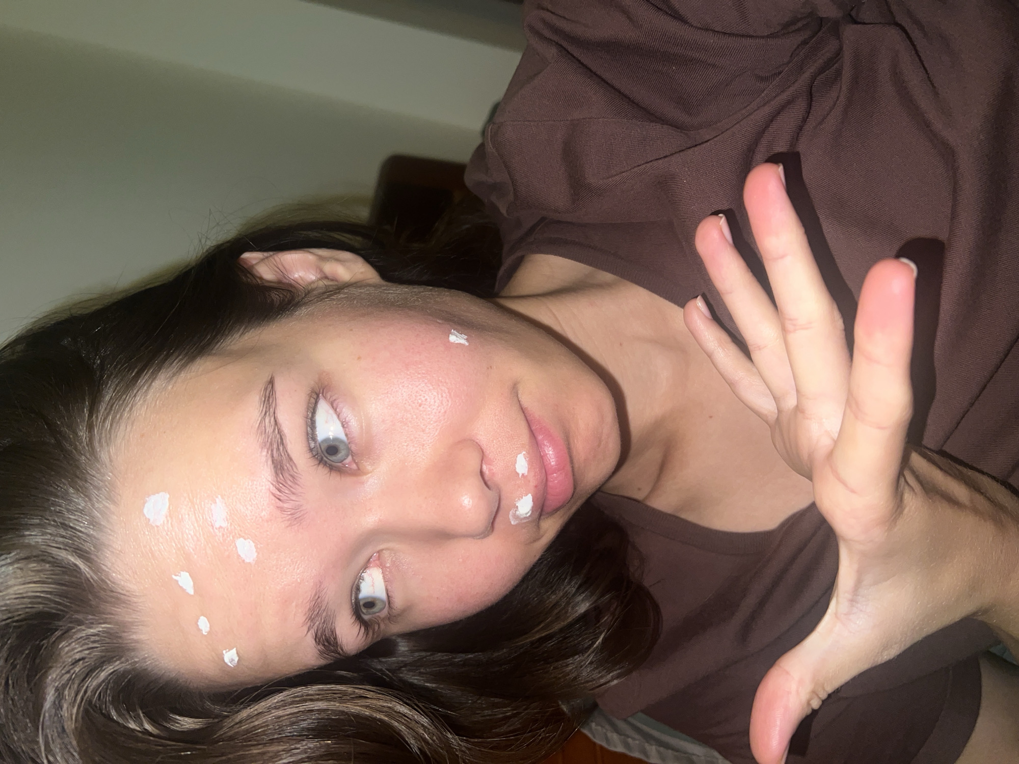 A woman with dark hair and blue eyes lies at an angle in an indoor environment. She is wearing a brown t-shirt. Several small white dots of cream are visible on her forehead and above her lip. Her right hand is raised with fingers spread. The location is Nam Nha Trang Ward, Vietnam.
