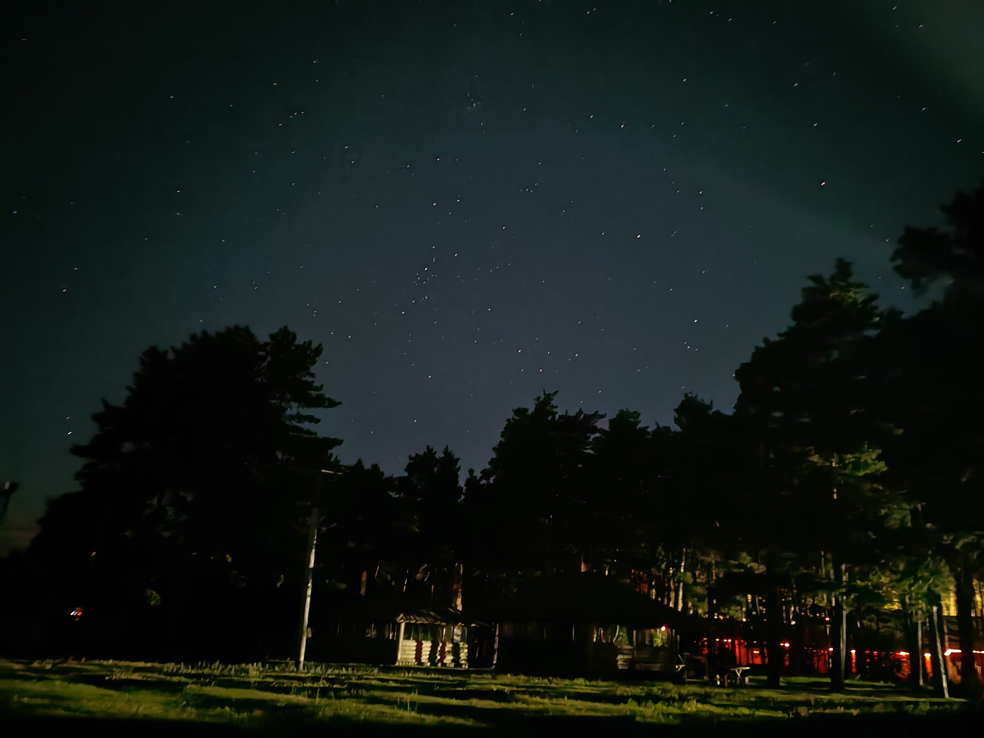 This media file captures a nighttime scene of a forest clearing with a rustic wooden structure. The sky above is dark and dotted with numerous stars, suggesting a clear night with minimal light pollution. Silhouetted trees frame the scene, their dark forms contrasting with the faint light illuminating the clearing.
The central subject is a long, low wooden building with a covered porch. It appears to be a communal or recreational area, possibly a picnic shelter or a simple cabin. Several smaller structures, perhaps outhouses or storage sheds, are clustered near the main building. The building's interior is dimly lit, with some warm-toned lights visible from within, casting a soft glow on the surrounding area. Red lights, possibly string lights or lanterns, are also visible strung between trees in the background, adding a festive or atmospheric touch.
In the foreground, a grassy area is illuminated by ambient light, with some patches appearing brighter than others, perhaps due to moonlight or artificial lighting. A single, tall pole stands on the left side of the clearing, its purpose unclear.
The scene evokes a sense of tranquility and remoteness, perfect for stargazing or a quiet evening outdoors. The overall impression is one of a peaceful, natural setting illuminated by the celestial bodies and subtle man-made lights. No people are visible, and there is no visible text in the image. The time of day is clearly night.