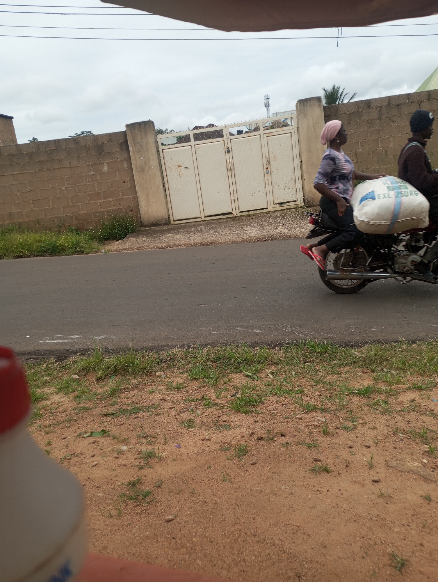 The image depicts a street scene in Jos, Nigeria. In the mid-ground, two individuals are riding a motorcycle. The person in the front, a woman wearing a pink headscarf and a patterned shirt, is facing forward, her left hand resting on a large, white sack secured to the back of the motorcycle. The sack appears to contain grain or similar produce and has text printed on it, including "EXL 250 KG". The person behind her, wearing a dark jacket and a black cap, is partially visible. The motorcycle is moving on a paved road. In the background, a brick wall with a cream-colored gate featuring decorative metalwork runs along the side of the road. Overcast skies are visible above. The foreground shows a dirt path with sparse green grass and some reddish-brown soil, with a blurry object, possibly a container, partially in frame at the bottom left.