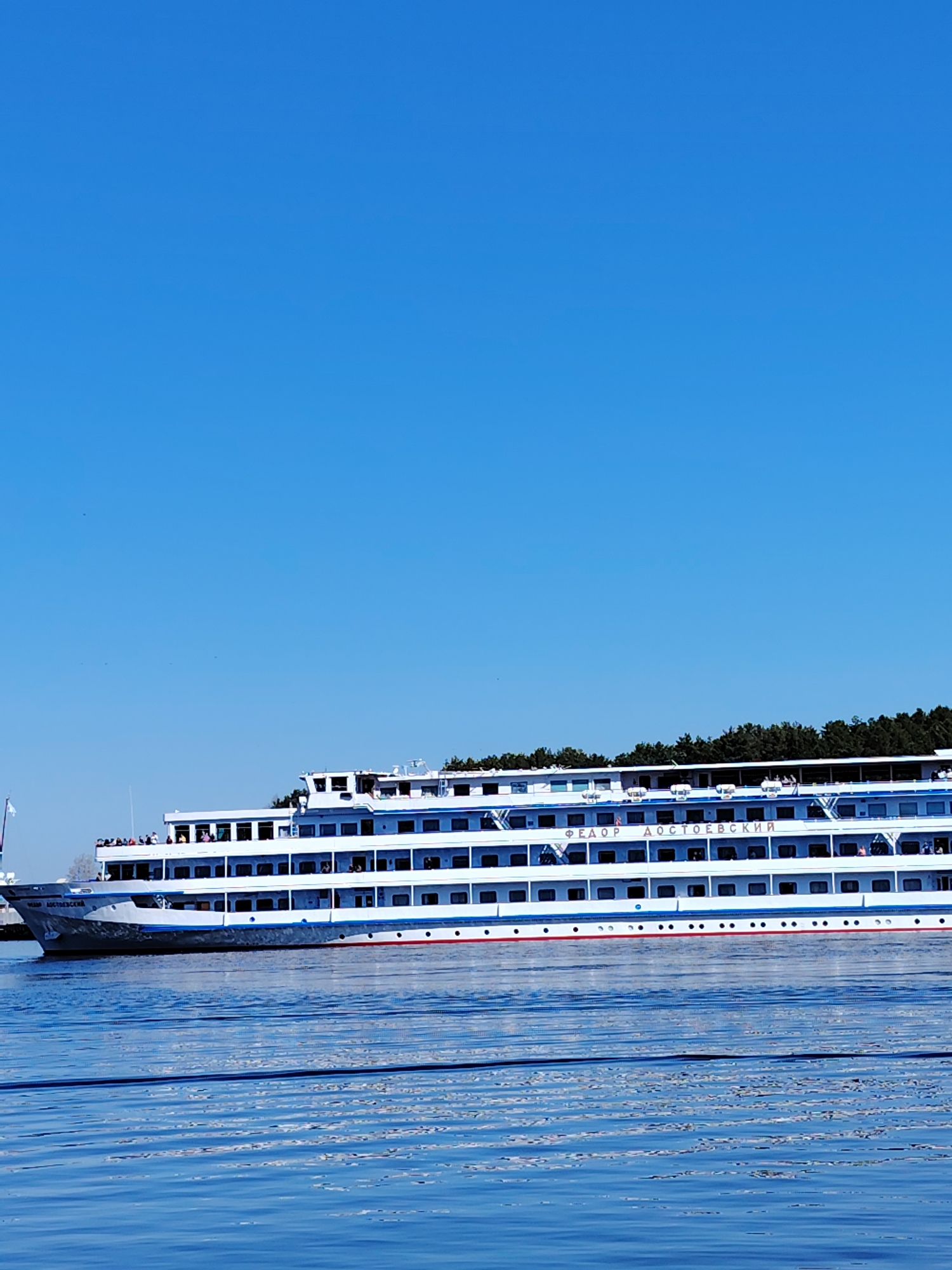 Image depicts the river ship "Fyodor Dostoevsky" on the water, likely in the city of Chaikovsky, Russia. The ship is white with blue stripes and red accents. It is multi-tiered and appears to be carrying passengers. The sky is a clear blue. The water has a rippled texture.