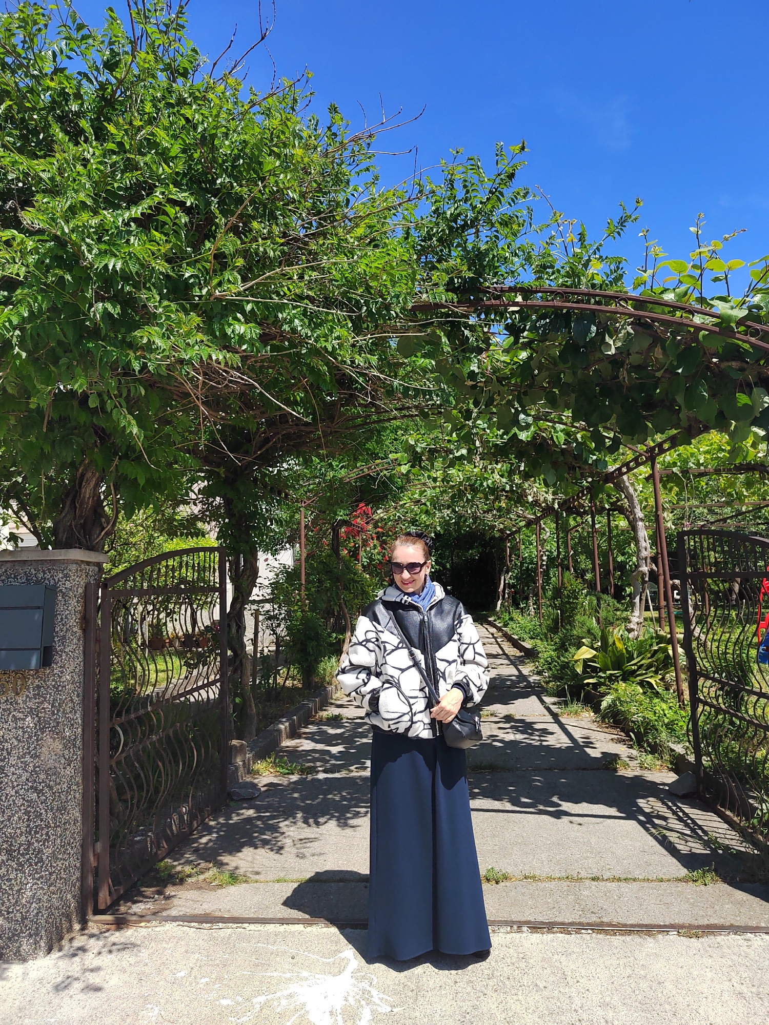 A woman stands centrally on a concrete pathway under a lush green pergola in Podgorica, Montenegro. She is wearing a dark blue full-length skirt or dress, a white and black geometric-patterned jacket, and sunglasses. A light blue garment is visible beneath her jacket, and she holds a black shoulder bag. Her expression is neutral as she looks towards the camera.
The pathway, marked with strong shadows from the overhead foliage, leads into the background, where more greenery and structures are visible. The pergola structure is made of dark metal arches heavily covered with vibrant green leaves and vines, creating a natural canopy. To the left of the woman, an ornate dark metal gate with a wavy design is visible, attached to a gray and black speckled stone pillar. A dark rectangular panel on the pillar clearly shows the number "10".
The scene is brightly lit by natural sunlight, with a clear, vivid blue sky visible above the dense foliage, suggesting it is midday or early afternoon on a sunny day. The overall setting is an inviting outdoor garden or pathway, characterized by abundant plant life and traditional architectural elements.