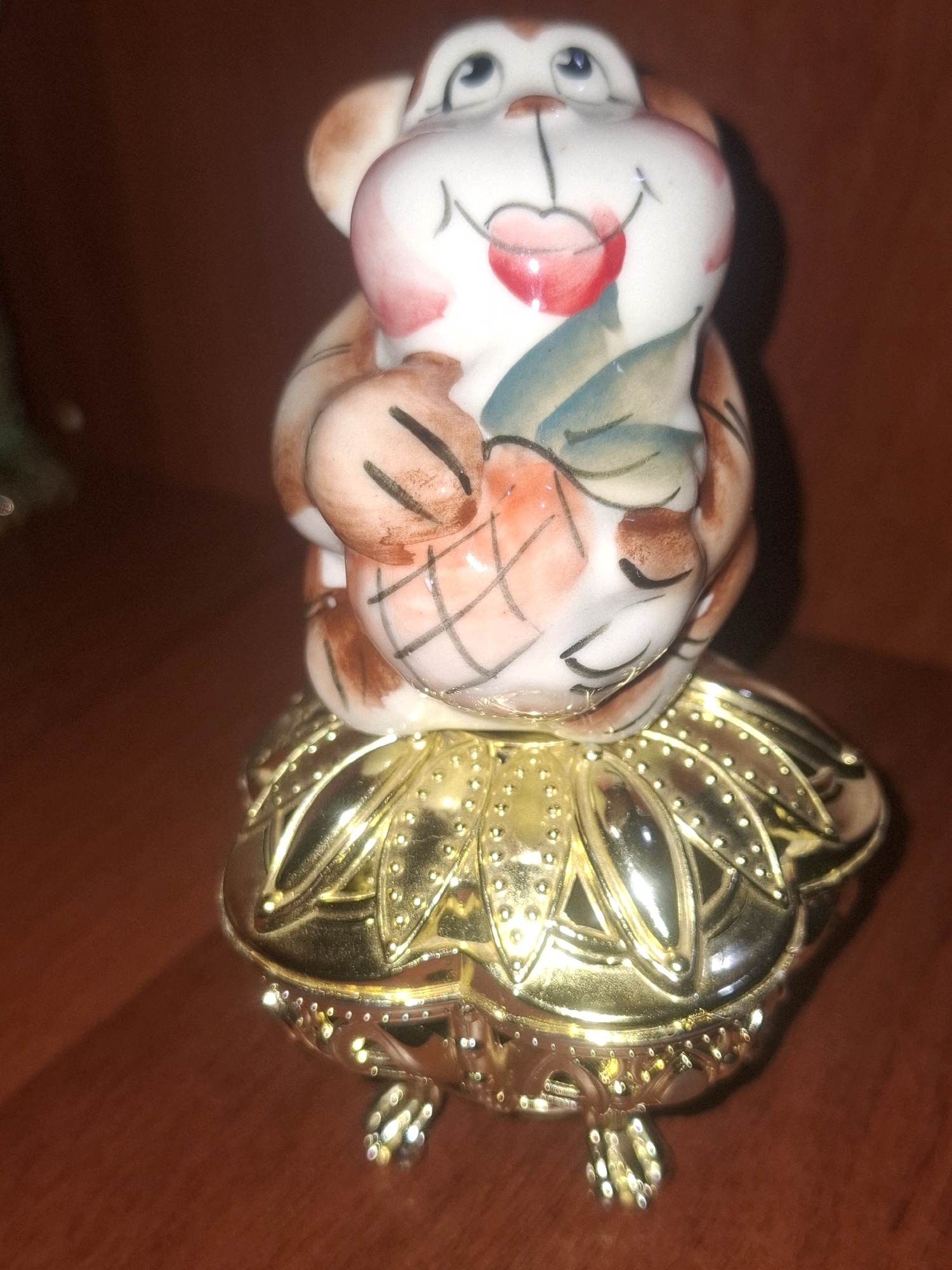 A ceramic figurine of a smiling monkey holding what appears to be a pineapple or fruit sits atop a decorative gold-colored base with leaf-like embellishments and small legs. The figurine is predominantly white with pink blush on its cheeks, brown accents on its limbs and ears, and a red heart-shaped mouth. Blue and green leaves are painted on the fruit it holds. The background is a dark brown, smooth surface, likely a wooden table. The overall lighting is dim, creating some shadows. The location is not determinable from the image.