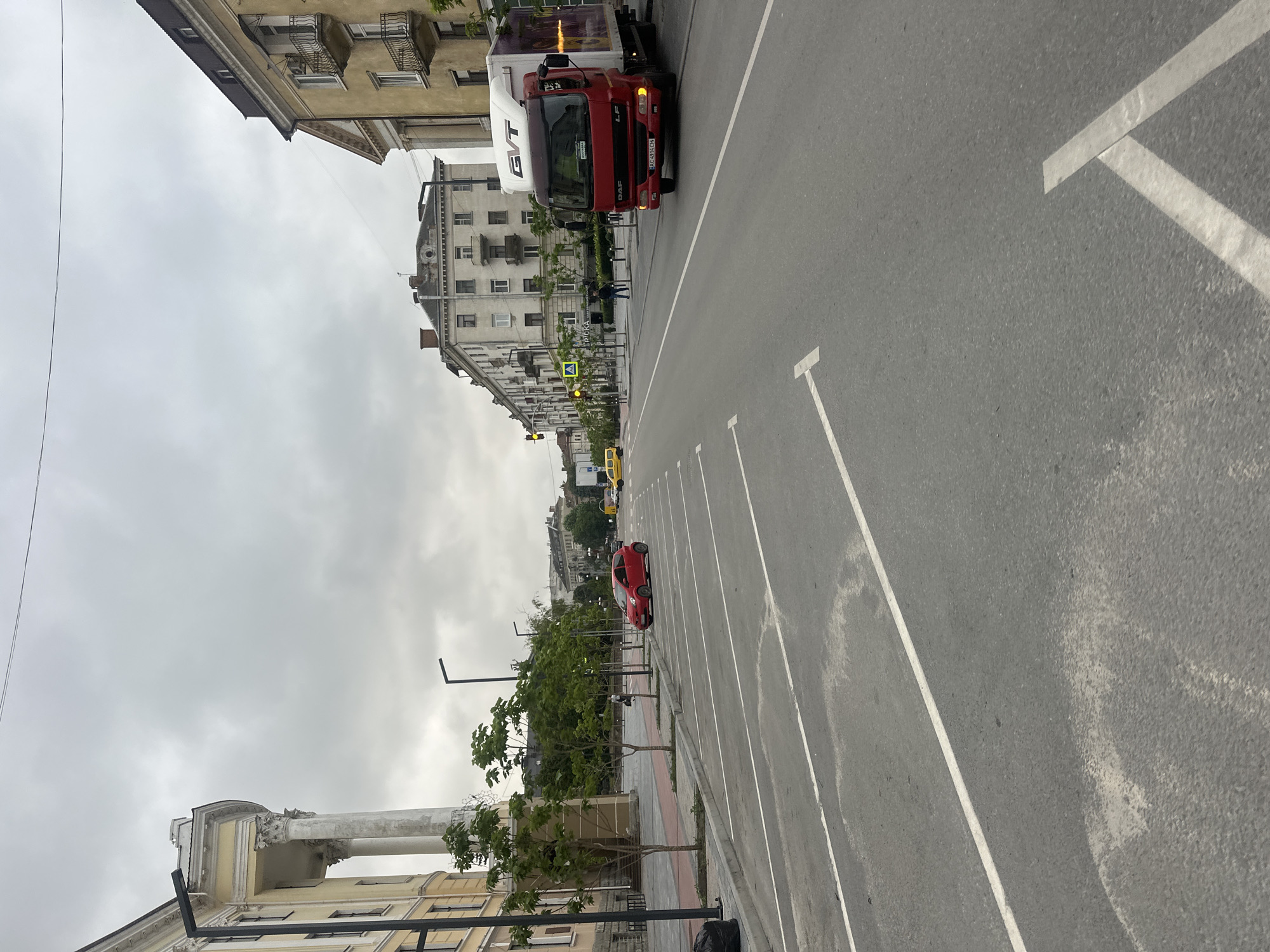 The image depicts a street scene in Ternopil, Ukraine, on an overcast day. In the foreground, a paved parking lot with white diagonal and straight lines is visible. A red car is parked on the left side of the street. Across the street, a red DAF truck with the logo "GVT" is driving towards the viewer, its license plate readable as AC 4914 CH. On the left side of the street, a large building with classical architecture, featuring columns and a light yellow facade, is prominent. Several young trees are planted along the sidewalk. Further down the street, multistory residential buildings line both sides. Traffic lights are visible, indicating intersections. A yellow taxi is seen in the distance on the left. The sky is cloudy, suggesting an overcast or possibly rainy day. The overall impression is that of a typical urban street with a mix of residential and commercial buildings, parked cars, and moving traffic.