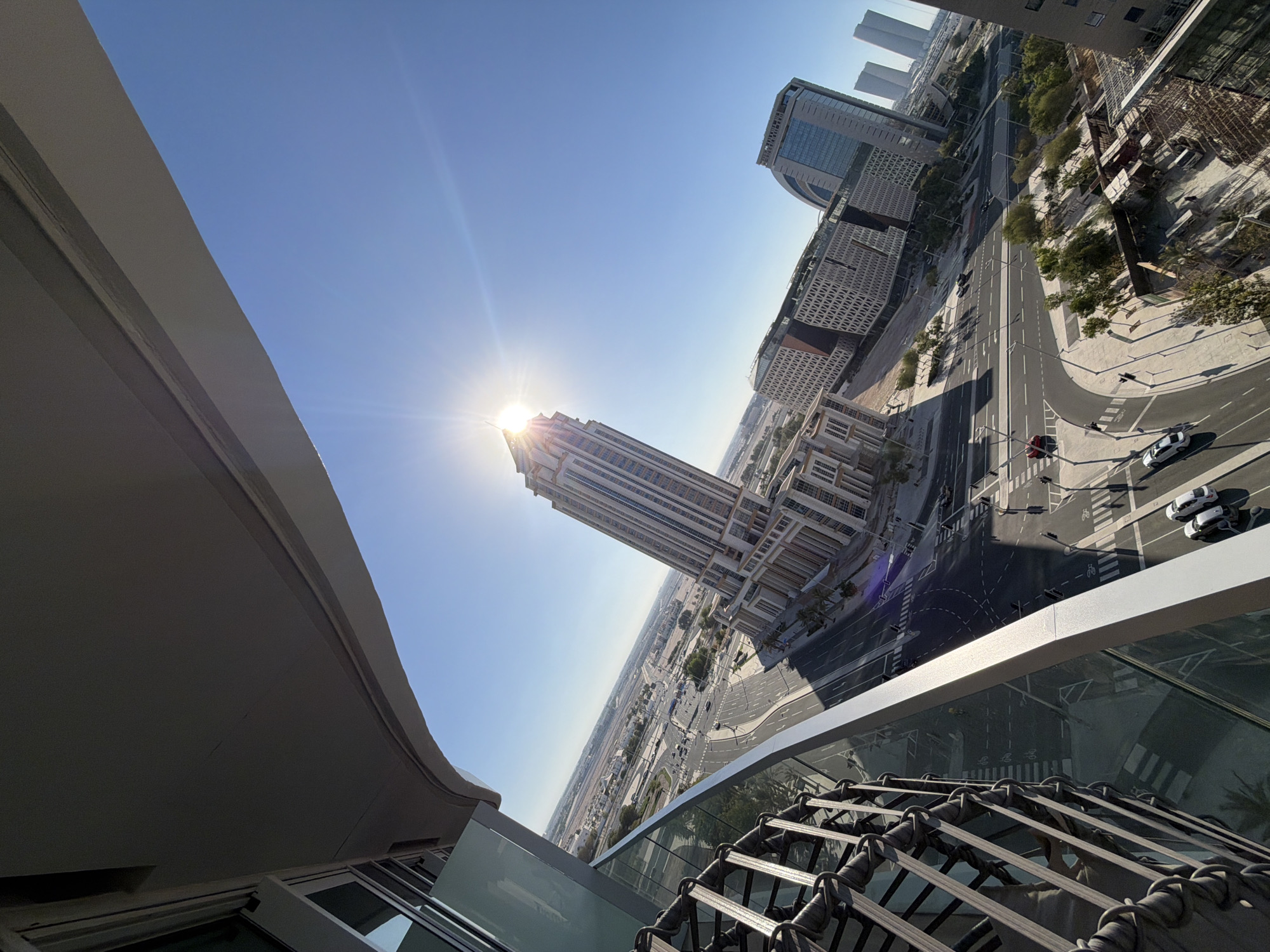 The image is an upward-angled, wide-angle shot of a city skyline under a clear blue sky. The sun is bright and positioned high, creating a lens flare that obscures some details. In the foreground, the edge of a building or balcony is visible, with a curved, gray architectural element dominating the left side of the frame. Below this, a glass railing and a decorative metal structure with a woven pattern are visible.
The main focus of the image is the urban landscape. Several modern skyscrapers of varying heights and architectural styles are present. One tall, slender building with a repeating pattern of windows stands out, and to its right, a wider building with a distinctive perforated facade is prominent. Further in the distance, other high-rise structures and a sprawling urban environment extend towards the horizon.
A multi-lane road curves through the city, with a few cars visible, including two white cars parked on the side of the road. Traffic lights and pedestrian crossings are also apparent. Greenery, in the form of trees, is interspersed throughout the urban setting. The overall scene suggests a bright, sunny day, likely mid-morning or early afternoon given the sun's position. The lack of clouds and the strong sunlight indicate clear weather. The architectural style and density of buildings point to a modern, well-developed city. No people are clearly discernible in the image, and no text is visible.