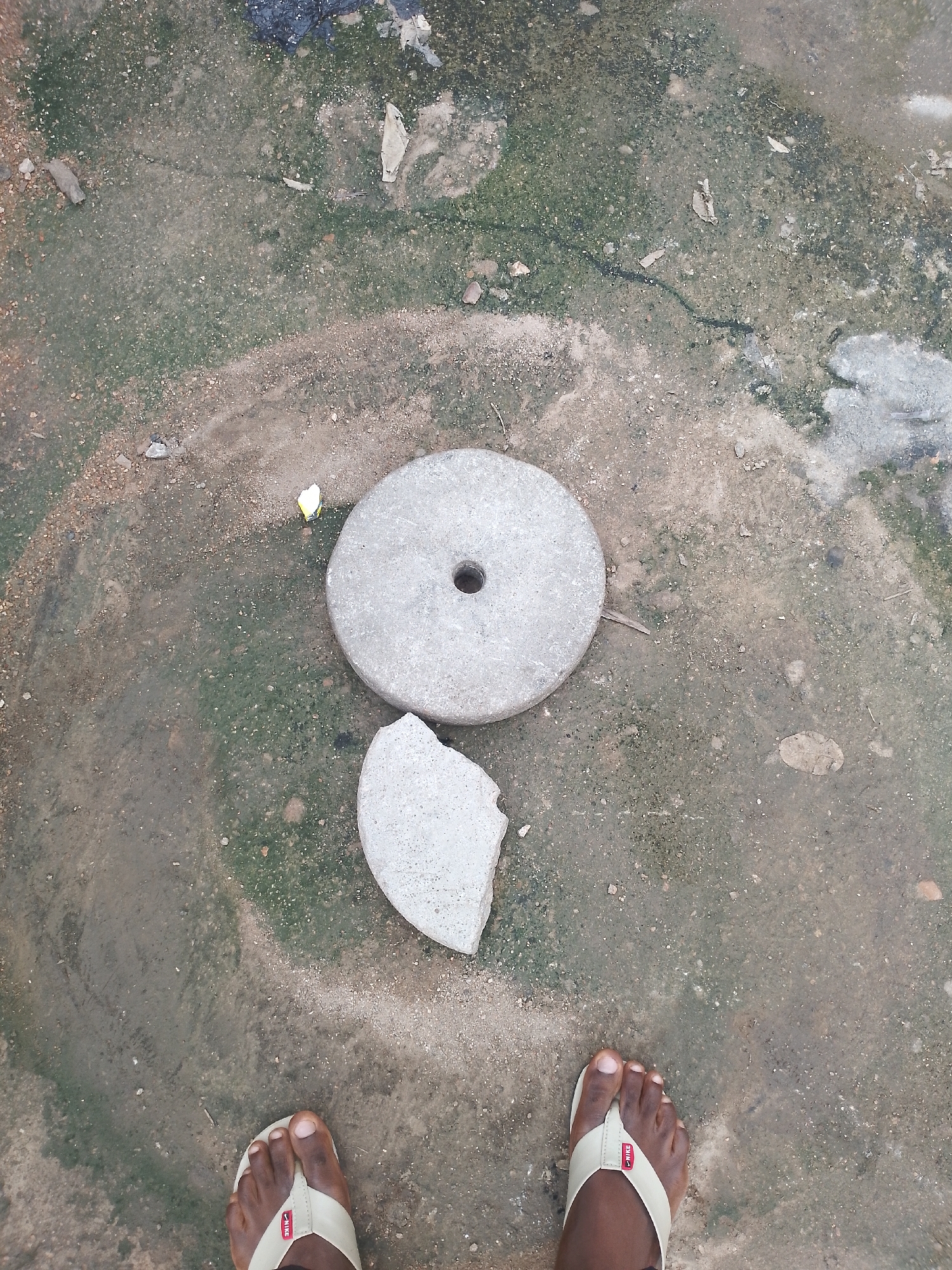 A pair of bare feet wearing light-colored flip-flops are visible at the bottom of the frame, positioned in the center. The feet are standing on a ground surface that appears to be a mixture of dirt, gravel, and patches of green moss. In the middle ground, there are two roughly circular stone objects. The larger one is intact and has a hole in the center, resembling a millstone. The smaller stone object is broken and appears to be a fragment of a similar stone. There is also a small, bright yellow piece of debris visible to the left of the intact stone. The ground is uneven and shows signs of wear and tear, with some cracks and scattered debris. The overall environment suggests an outdoor, possibly unpaved area.