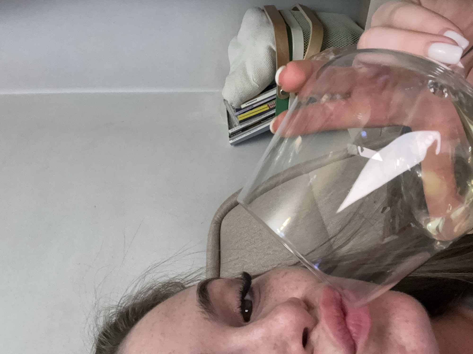 A woman holds a glass filled with a light-colored liquid near her face. The background includes a light gray wall and a collection of items stacked together, including a white cloth and magazines with colorful covers. There is no information present that indicates this is in the city of Plovdiv, Bulgaria.