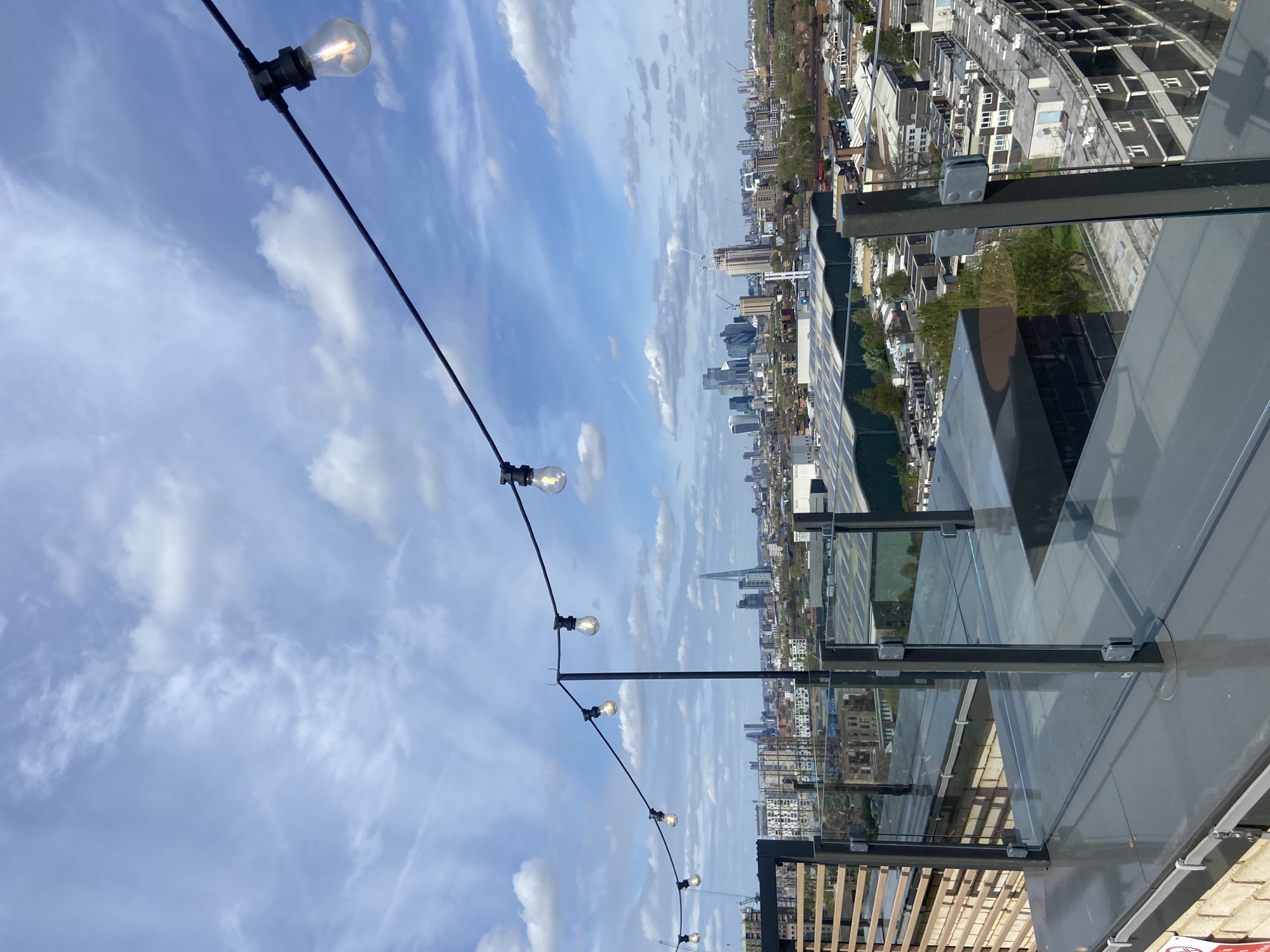 The image is a view from a high vantage point, likely a rooftop in London, United Kingdom. A string of bare light bulbs stretches across the sky, suspended from a wire. Below, the London skyline is visible, including the Shard skyscraper. The cityscape includes a mix of residential and commercial buildings, interspersed with greenery and what appears to be a waterway. The image is taken from behind a glass barrier, a design element of the rooftop structure. The sky is blue with wisps of white clouds.