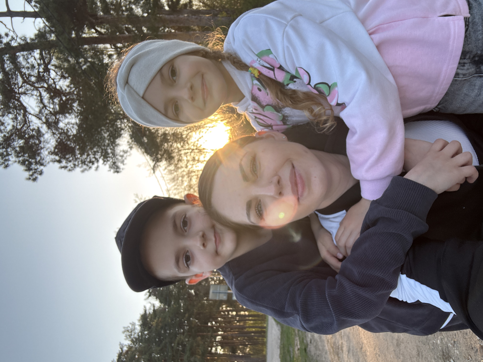 A woman is posing outdoors with two children, likely her children. The older child is on top, wearing a white head covering and a pink and white sweatshirt. The younger child is below her, wearing a black baseball cap and a navy blue sweatshirt. The woman is between the two children. The setting is in Раздолье, Russia, in an outdoor environment, possibly near a park, with trees visible in the background. The sun is shining.